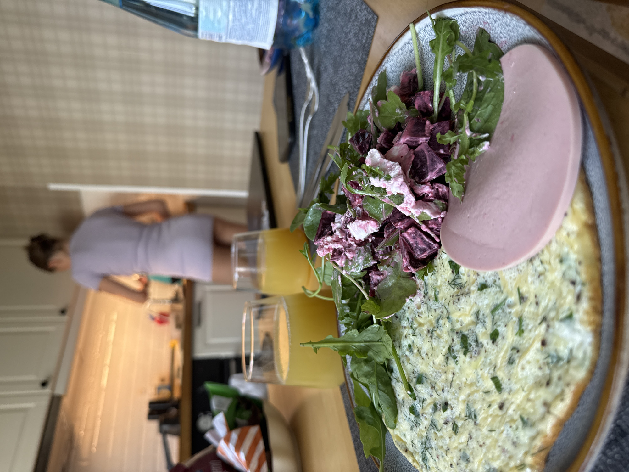In a bright, modern kitchen area that appears to be in Moscow, Russia, a woman with her hair pulled back is seen from behind, wearing a light purple dress and standing at the counter, seemingly preparing food. The foreground is dominated by a table laid out for breakfast. A plate holds a fluffy green omelet, a slice of pink bologna, and a vibrant salad of cubed beets, greens, and a creamy pink dressing. Two glasses of orange juice and a bottle of water are also on the table, alongside a fork and knife. The overall impression is one of a home cooked meal in a contemporary domestic setting.