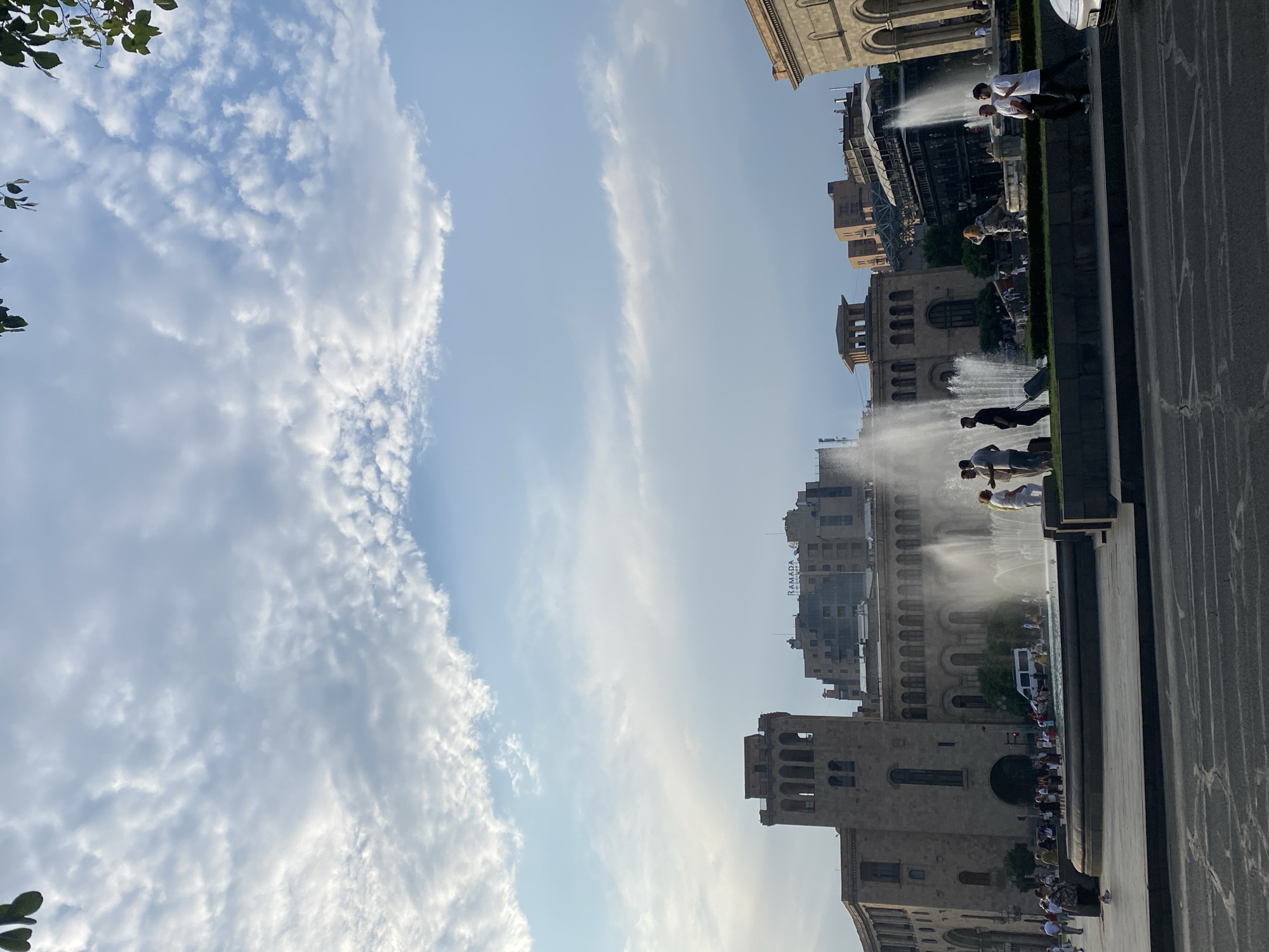 The image depicts Republic Square in Yerevan, Armenia, a bustling public space featuring a prominent fountain and several significant architectural structures. The sky above is a mixture of blue and scattered white clouds, suggesting it is daytime, possibly late afternoon or early evening given the lighting.
In the foreground, a wide paved area leads to a tiered fountain with multiple jets of water actively spraying. Several people are visible around the fountain, some standing and conversing, others walking. One individual is seen pulling a wheeled suitcase, suggesting they might be traveling. A white van is parked near the fountain on a road behind it.
Behind the fountain, a large, imposing building with a classical architectural style dominates the center of the frame. This is likely the National Gallery of Armenia or the History Museum of Armenia, both located on Republic Square. To the right and slightly behind this building, another multi-story building with the "RAMADA" sign is visible, indicating a hotel. Further to the right, another building with arched windows and classical elements can be seen.
The scene is alive with activity, showing people enjoying the public space, with the fountain serving as a central attraction. The overall impression is one of an urban environment, a meeting point for residents and potentially tourists, bathed in natural light.