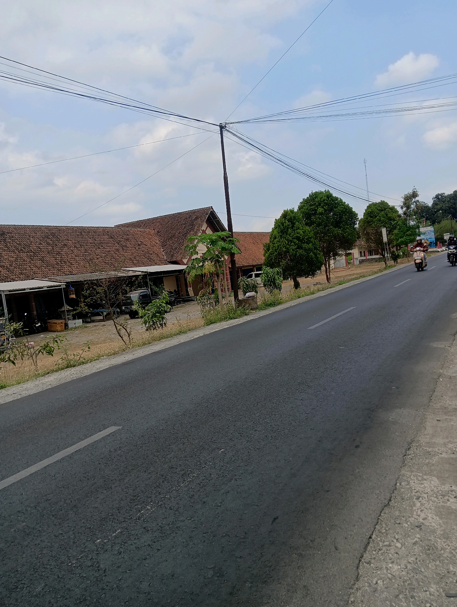 The image depicts a roadside scene in Sokaraja, Indonesia. A paved asphalt road runs diagonally across the frame from the bottom left to the upper right. The road has a white dashed line marking the center. To the left of the road, a long, single-story building with a tiled roof is visible. This building appears to be a commercial establishment, with open-fronted sections that house parked motorcycles and what seems to be a small truck. There are also some plants and small trees growing in front of the building.
On the right side of the road, the terrain slopes slightly upwards. Several mature green trees stand along this side. Further down the road, two individuals on scooters are traveling away from the viewer. The scooter closer to the viewer has a light-colored body and a rider wearing a helmet and a dark jacket. The second scooter, further away, is darker in color. The sky above is partly cloudy with a mix of blue sky and white clouds. Overhead, multiple power lines are strung across the scene, supported by poles. The overall impression is a rural or semi-urban street in Indonesia.