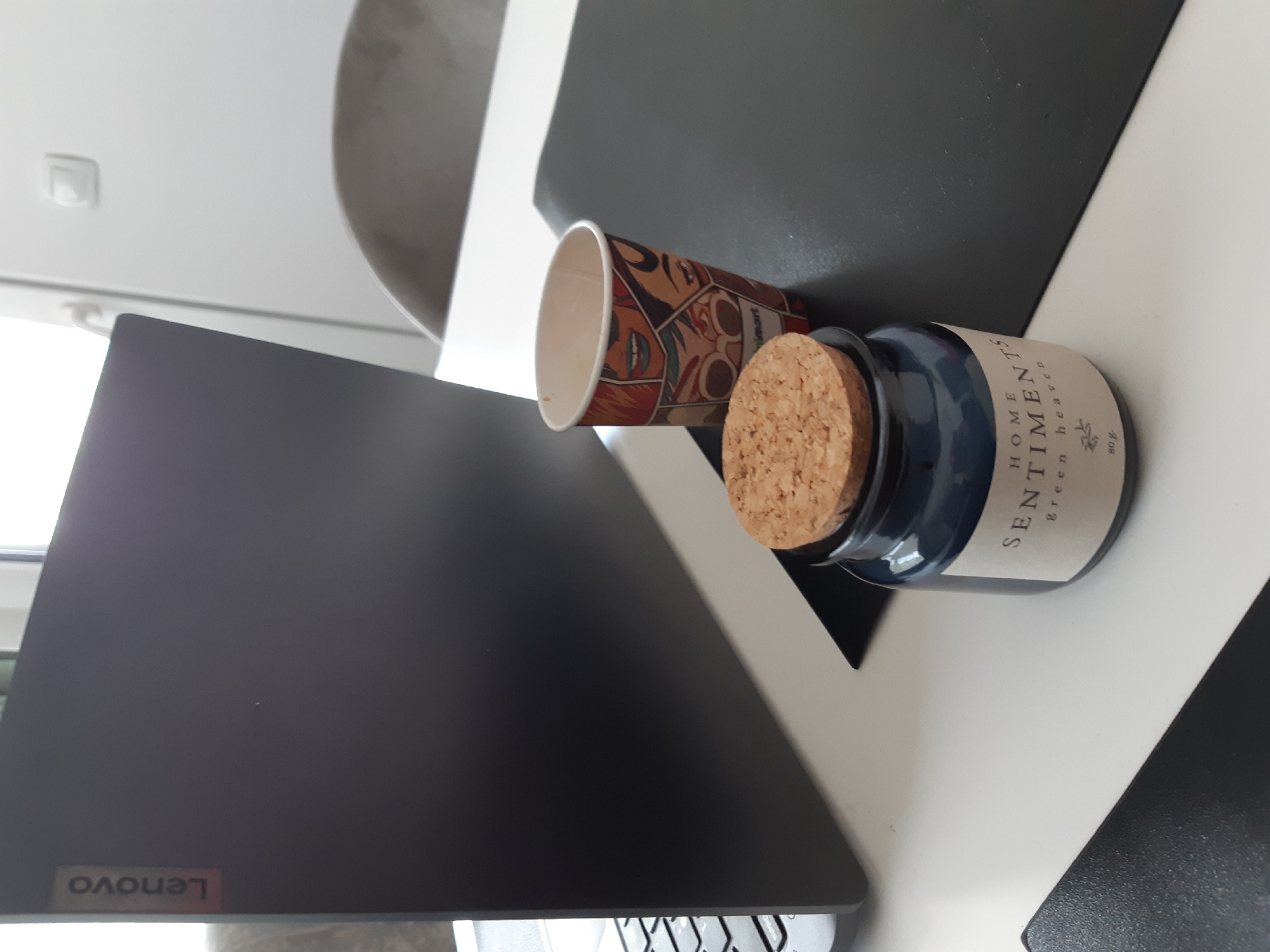 The image shows a close-up view of a desk or table setting. A Lenovo laptop occupies the lower left of the frame. Adjacent to the laptop are a disposable paper cup with a colorful comic book-style design and a small, blue glass jar with a cork stopper. The jar is labeled "Sentiments Home" and "Green Heaven 80g." The surface appears to be a white and black desk or table, likely indoors, as evidenced by the ceiling light fixture in the upper left. The presence of the laptop, coffee cup, and scented jar suggests a workspace or personal area. The location in Plovdiv, Bulgaria is not discernible from the image.