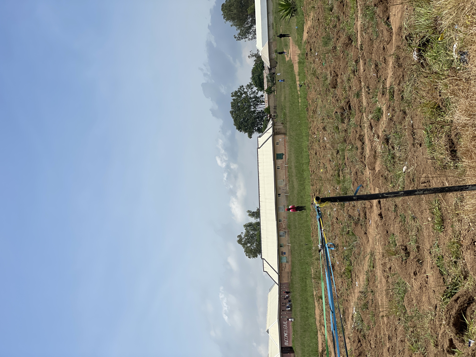 The image depicts a view of buildings in Durumi, Nigeria, under a partly cloudy sky. In the foreground, a construction area is visible with disturbed earth and some yellow and blue construction ribbons attached to a black pole. Beyond this, a grassy field separates the construction site from several low-rise, light-colored buildings with white roofs. Some of these buildings have graffiti or writing on their walls, including the words "INCONCLUSIVE" and other illegible marks. A few people are scattered around the buildings and in the field, with one person in a red vest standing in the middle ground. Trees are visible behind and between the buildings. The overall impression is of an unfinished or developing area.
