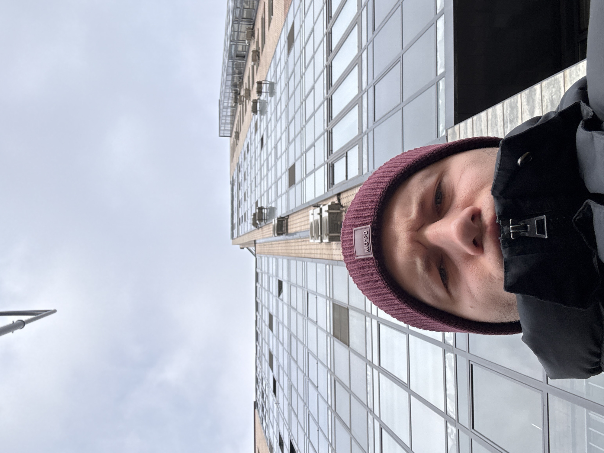 A man wearing a maroon beanie and a dark jacket is positioned in the lower right corner of the image. His face is partially visible, from the eyebrows to the nose. A tall building with numerous windows stretches upwards to the right of the frame. The sky, visible on the left side, is overcast with a greyish-white hue. A street lamp is visible in the bottom left corner. The location is likely Krasnoyarsk, Russia, based on the man's attire and architectural style of the building.