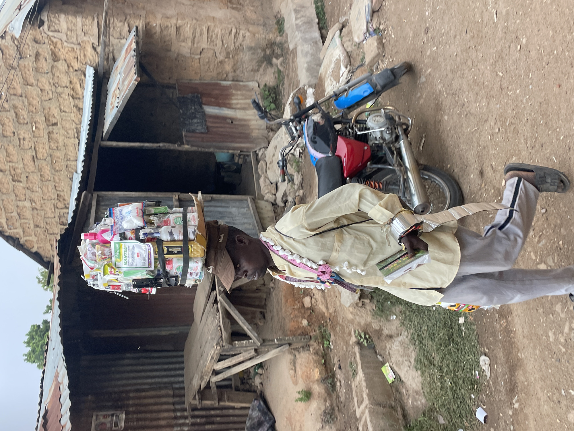 A man, likely a street vendor, stands outdoors, carrying a large, diverse stack of goods on his head. He wears a light-colored long-sleeved shirt, light grey trousers with a dark stripe, and dark sandals. A light brown cap covers his head beneath the carried items. Around his neck, he has a pink measuring tape and a necklace adorned with white pom-poms, from which a pair of scissors is suspended.
The items on his head form a tightly packed mound, secured with straps. They appear to be various packaged products, including bottles, boxes, and pouches. Visible text on some packages includes "MANTEL" (on a prominent green packet) and "BLUE DIAMOND." He holds a small, rectangular item in his left hand.
The background shows a dusty, unpaved area, typical of a village or town street. To his left, there is a building with a textured stone wall and corrugated metal sheets used for roofing and siding. To his right, a utility-style motorcycle, black and red with blue accents, is parked. The ground is a mix of dirt and scattered debris, with some sparse vegetation. The lighting suggests it is daytime, possibly with an overcast sky or soft sunlight. The scene is consistent with a market or street hawking environment in a location like Jos, Nigeria.