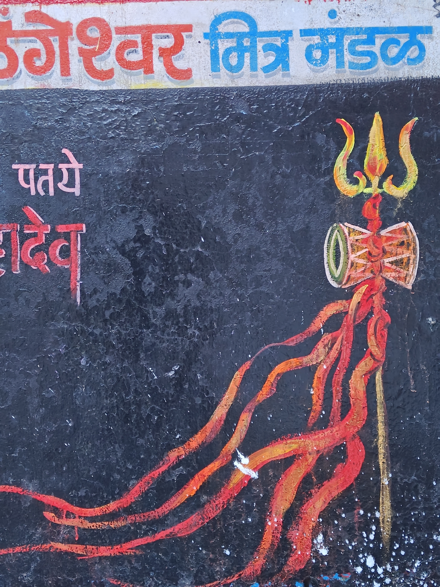 The image shows a rough, dark textured wall featuring a painted trident (trishul) above a drum (damaru), which are associated with the Hindu deity Shiva. The trident is predominantly yellow and orange, while the drum is a mix of red, orange, and green. Red and orange lines, resembling flames or energy, extend down from the drum. Above these depictions are words painted in red and blue, likely in a local Indian language, possibly Hindi or Marathi, which could represent the name of a local organization or association. To the left, more writing appears in pink and red. The specific location in Mumbai, India, cannot be determined from the visual information provided.