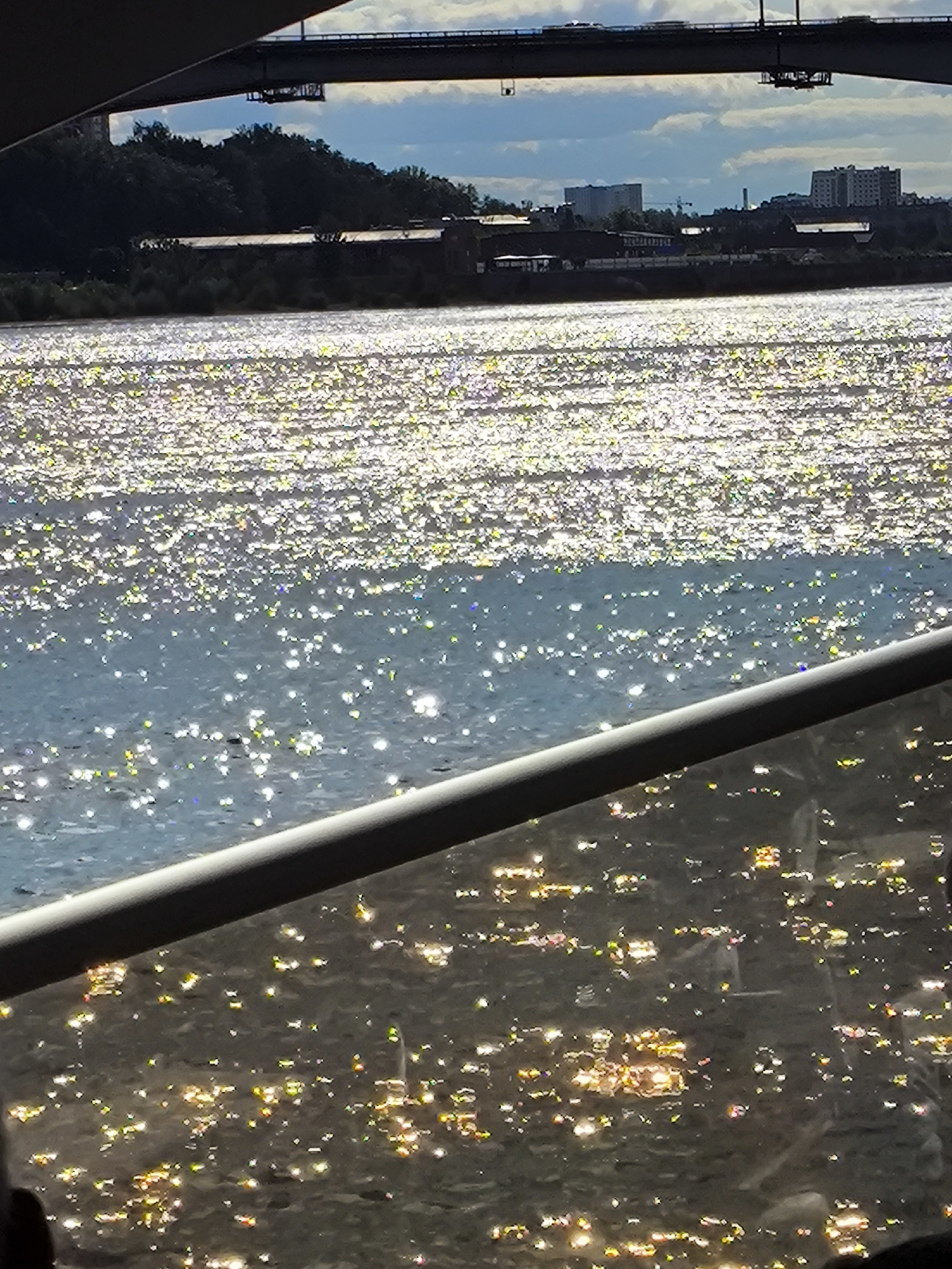 The image captures a shimmering body of water, likely a river, reflecting bright sunlight. In the background, a large bridge spans across the scene, with vehicles visible on its roadway. The far bank of the river features a tree-lined shore with buildings and structures dotted along the landscape. The foreground shows a dark railing, possibly from a boat or viewing platform, with reflections of lights on the water's surface, suggesting an urban setting at dusk or night. The scene evokes a sense of movement and light. This view is consistent with the Kama River embankment in the city of Perm, Russia, possibly near the Kama River Bridge. No people are clearly identifiable in the image.
