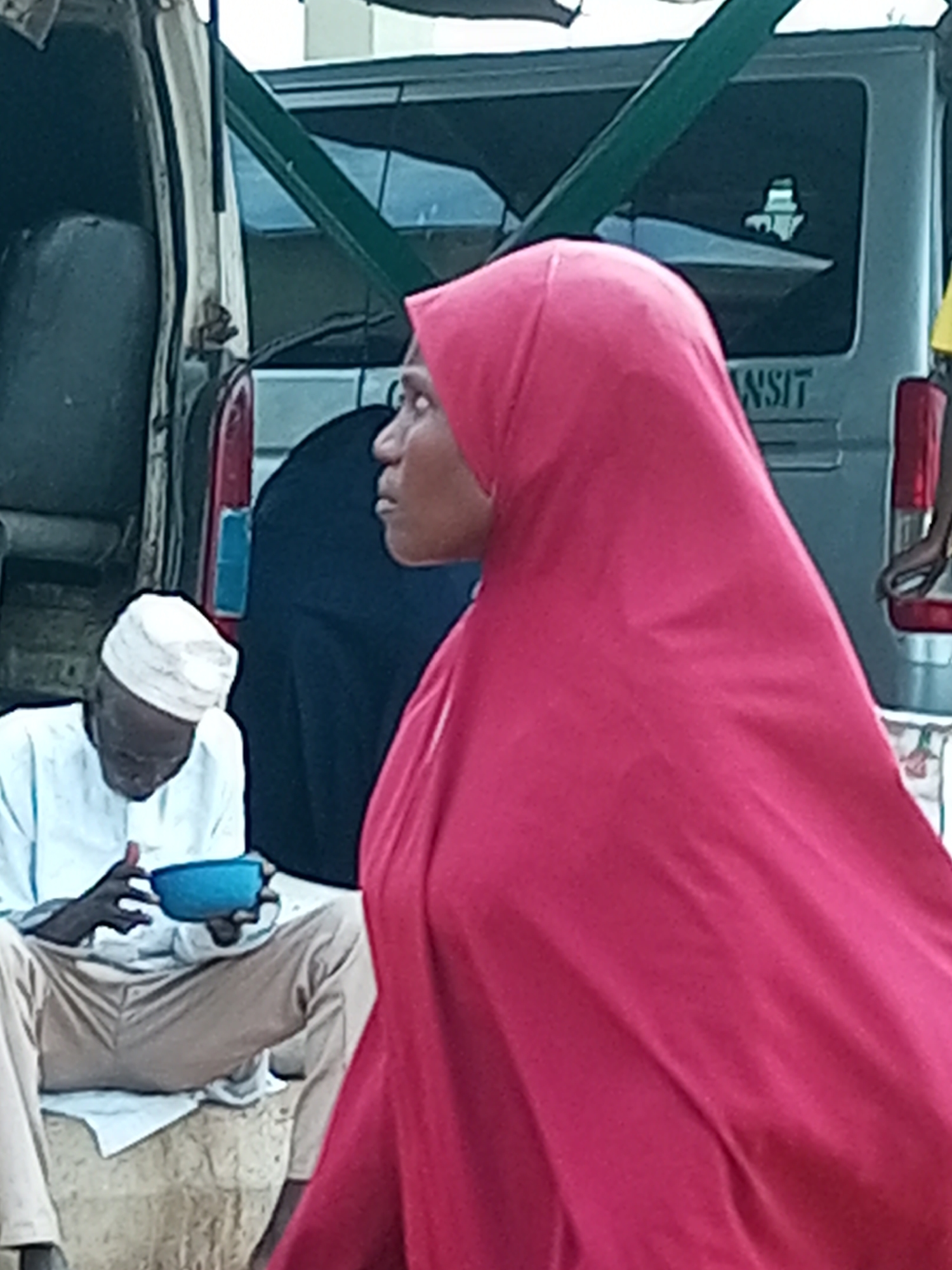 In Gombe, Nigeria, a person wearing a red hijab is captured in a profile view, facing left. To the left and slightly behind this individual, an elderly man wearing a white cap and a white robe is seated, holding a blue bowl in his hands. The background shows a vehicle, possibly a van, with a green frame across its rear window, and some lettering on the rear of the vehicle reads "NSIT". The scene appears to be outdoors, with a rough, textured surface beneath the seated man.