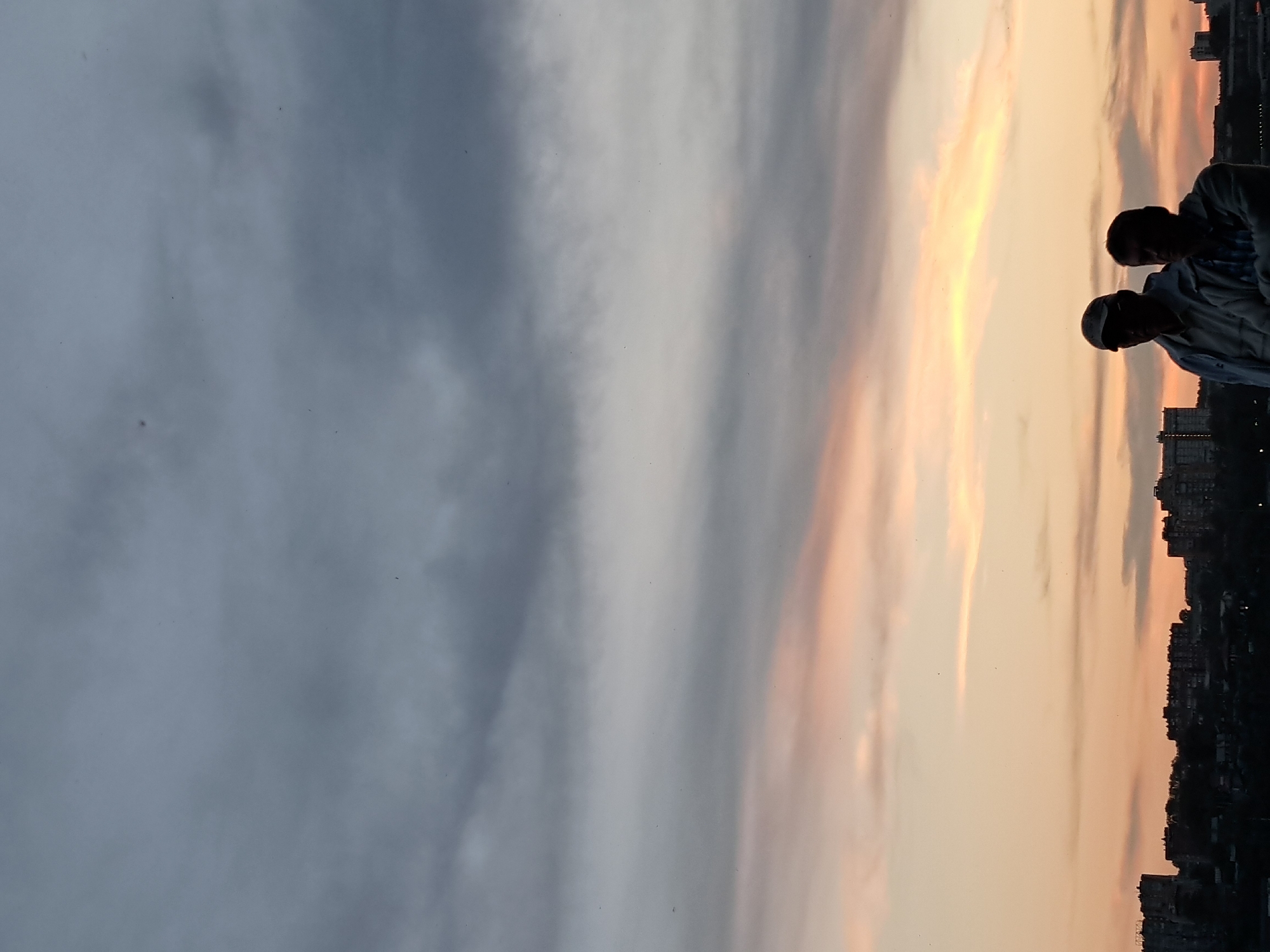 The image captures two men standing silhouetted against a dusky sky over the city of Irkutsk, Russia. The sky is a gradient of gray clouds at the top, transitioning to soft pink and orange hues near the horizon. The city skyline, also silhouetted, features apartment buildings and other structures. The man on the left wears a baseball cap and a light-colored jacket, while the man on the right is dressed in a plaid shirt and a jacket. They appear to be standing close together, possibly observing the sunset. The overall impression is one of quiet contemplation as day turns to night.