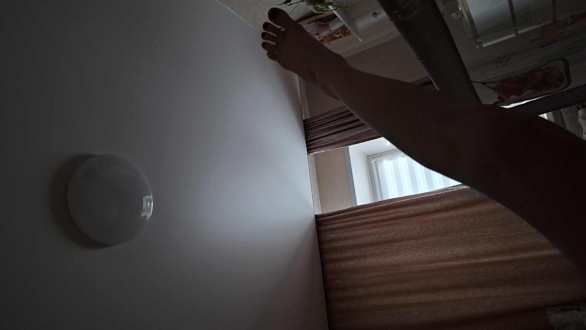 A view from below of a person's right leg extending upwards. The foot is visible with painted toenails. The room contains a white wall with a round light fixture, a window with white vertical blinds, and curtains. The location is Chaikovsky, Russia.