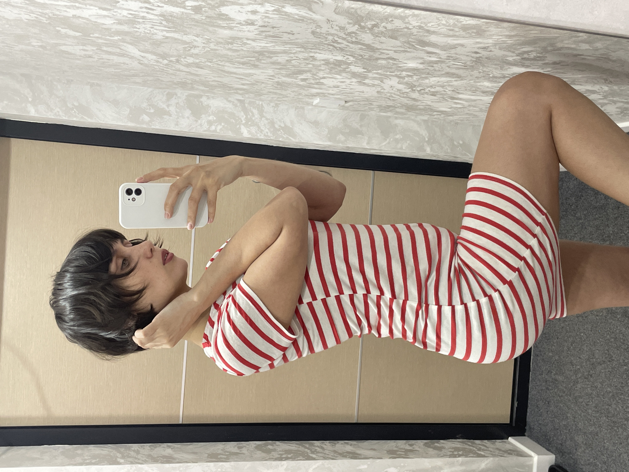 A woman is taking a selfie in a room. She is lying on her side, wearing a red and white striped dress. The walls are cream-colored with black trim. The floor is gray. A marble-patterned surface is visible in the upper portion of the image. The city of Izhevsk, Russia cannot be ascertained from this image.