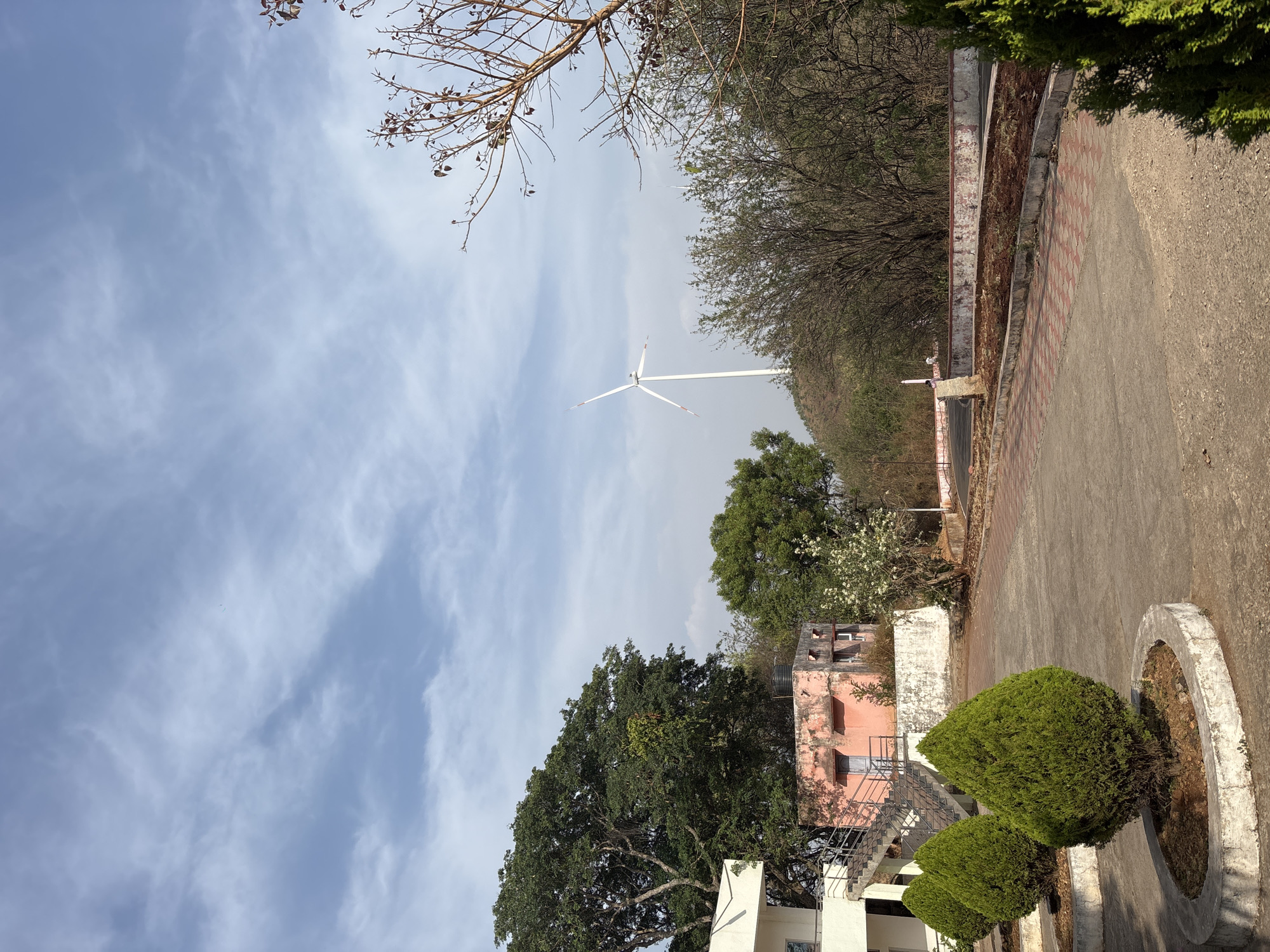 The image, taken in Hosapete, India, shows a wind turbine standing tall in the distance against a backdrop of a blue sky with scattered clouds. The turbine is the prominent feature in the mid-ground. The foreground features a paved road or pathway with a low wall or embankment on one side and some greenery, including trees and bushes. A light pink building with stairs is visible amongst the trees. The upper portion of the frame shows bare tree branches against the sky.