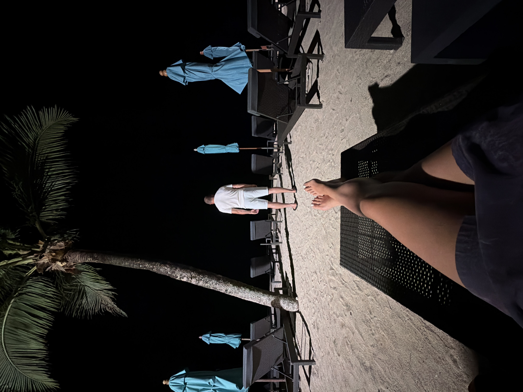 In Bang Muang, Thailand, a person lies on a beach lounger, their legs extended towards the foreground, casting a shadow on the sand. They are wearing dark shorts. Facing away from the camera, a man in a white t-shirt and light-colored shorts stands on the sandy beach. Behind him, several empty, dark-colored lounge chairs and closed turquoise beach umbrellas are arranged on the sand. A palm tree with its fronds illuminated by an unseen light source stands to the left of the frame, its trunk leaning slightly. The background is dark, indicating nighttime.