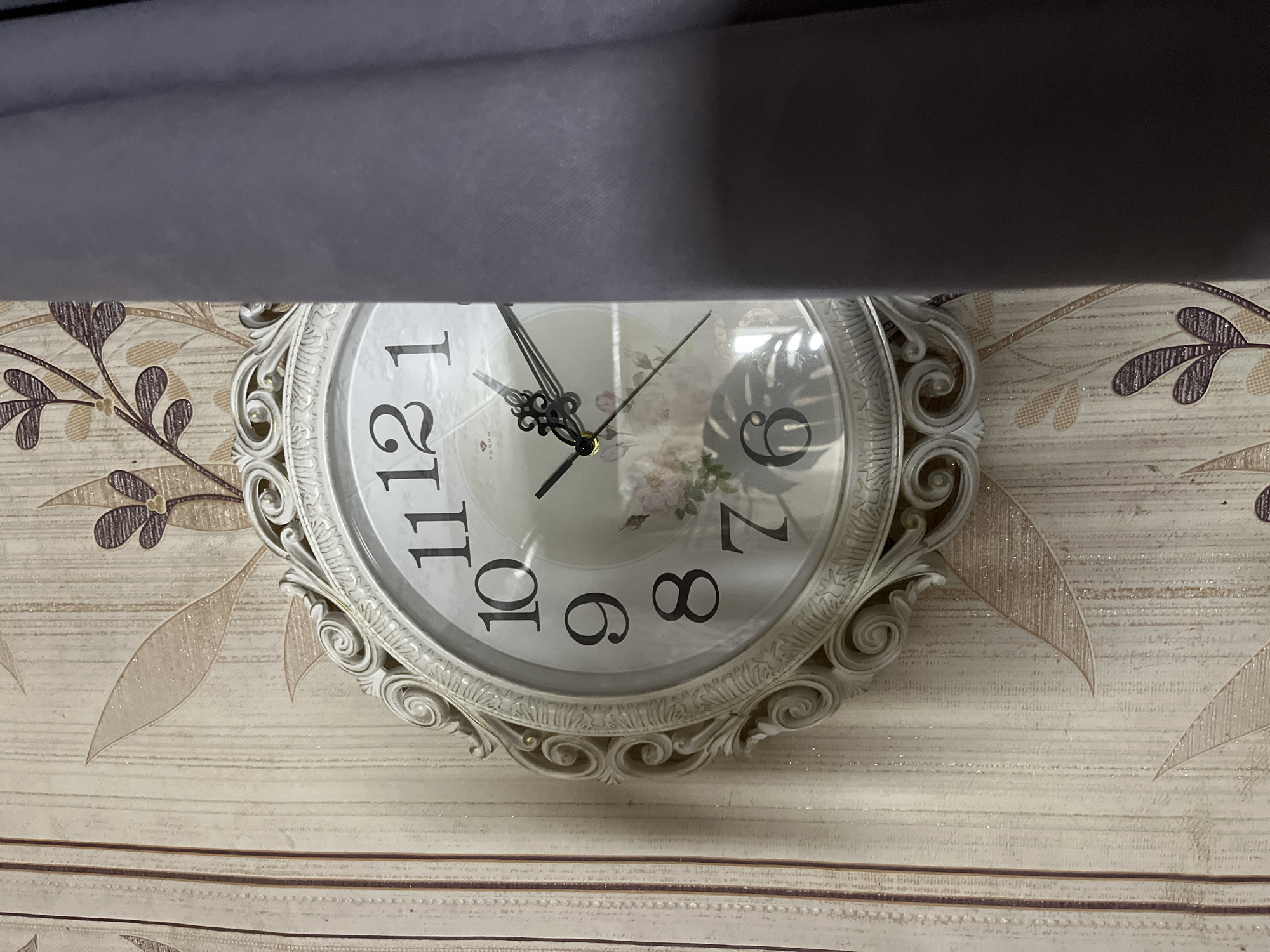 A close-up shot of a white ornate clock hanging on a wall with beige and brown floral wallpaper. A gray fabric, possibly a curtain or drapery, obscures the top part of the image. The clock's hands indicate a time around 11:25. The clock face has floral accents. The photo appears to be taken indoors. The location, Malyi Bukor, Russia, is not discernible from the image.