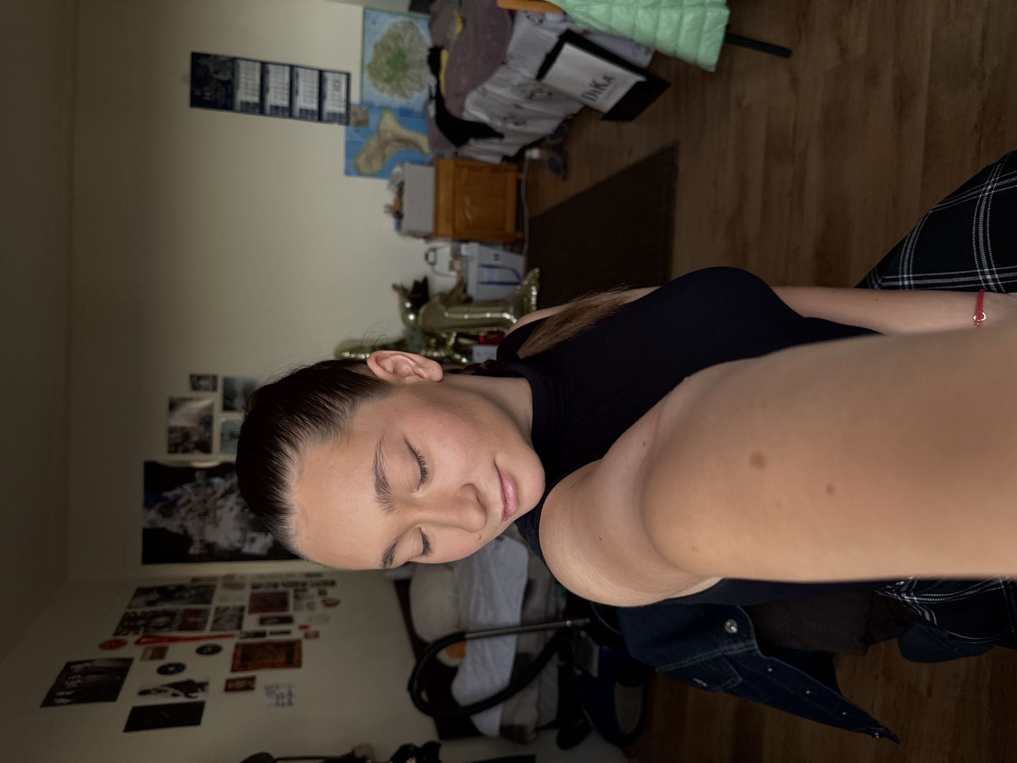 The image shows a young woman with her eyes closed, appearing to be in a relaxed or sleepy state. She is wearing a black top and a plaid skirt. A red bracelet is visible on her wrist. The setting appears to be a room, likely a bedroom, with posters and pictures on the wall. A wooden floor is visible in the background. There is some clutter on a bed or sofa behind her, including what looks like a green blanket and some pillows. A wooden nightstand with drawers is also visible. The lighting suggests it is likely indoors, possibly at night or with dim lighting. The overall mood is intimate and personal. The location is Sofia, Bulgaria. There is no visible text in the image.