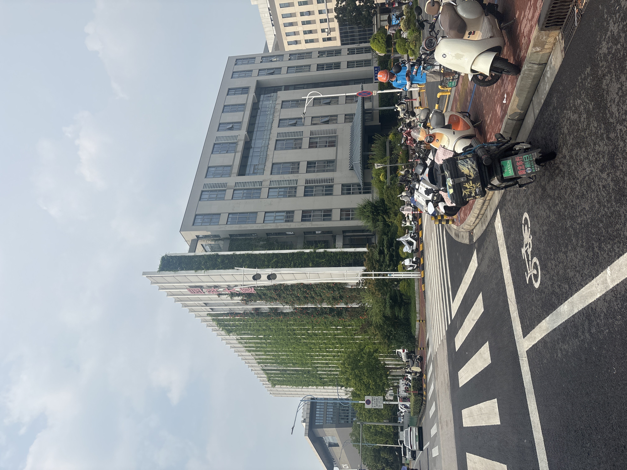 In a city scene in Suzhou Industrial Park, China, a modern building with a vertical green wall stands prominently. The building has a white facade with vertical white fins and is adorned with lush green plants cascading down its side. A portion of the building to the right is a grey structure with numerous windows. In front of the building, a street with a pedestrian crossing is visible. Several electric scooters and motorcycles are parked on the sidewalk. A person wearing a blue shirt and an orange helmet is standing near the parked vehicles, looking at their phone. The sky above is partly cloudy.