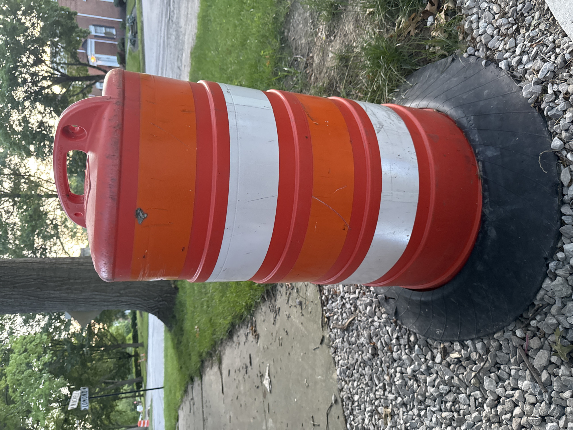 An orange and white striped traffic barrel stands on a base of gravel next to a sidewalk. The barrel has a red handle on top and is positioned on a black rubber mat. In the background, trees and residential buildings are visible, along with street signs indicating "WINCHELL" and "LOMOND." The setting appears to be a residential street in Shaker Heights, Ohio, United States of America. The lighting suggests it is either early morning or late afternoon.