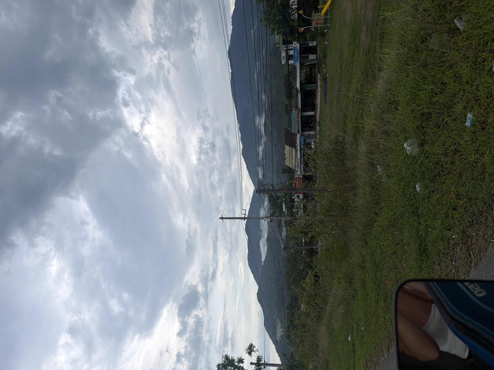 The image shows a view from a vehicle in Diên Thọ, Vietnam, looking towards a mountain range under a cloudy sky. In the foreground, tall grass lines the side of a road. Several utility poles with numerous electrical wires are visible, running across the landscape. In the mid-ground, a row of small buildings, likely shops or residences, sits at the base of the mountains. Some of these buildings have signs, and a yellow playground slide is visible to the right of the buildings. The mountains themselves are covered in dense green vegetation and partially obscured by low-lying clouds or mist, particularly in the valleys. The overall scene depicts a semi-rural environment with infrastructure and natural landscape elements. The reflection in the side mirror shows a person wearing a white tank top and a portion of a blue vehicle.