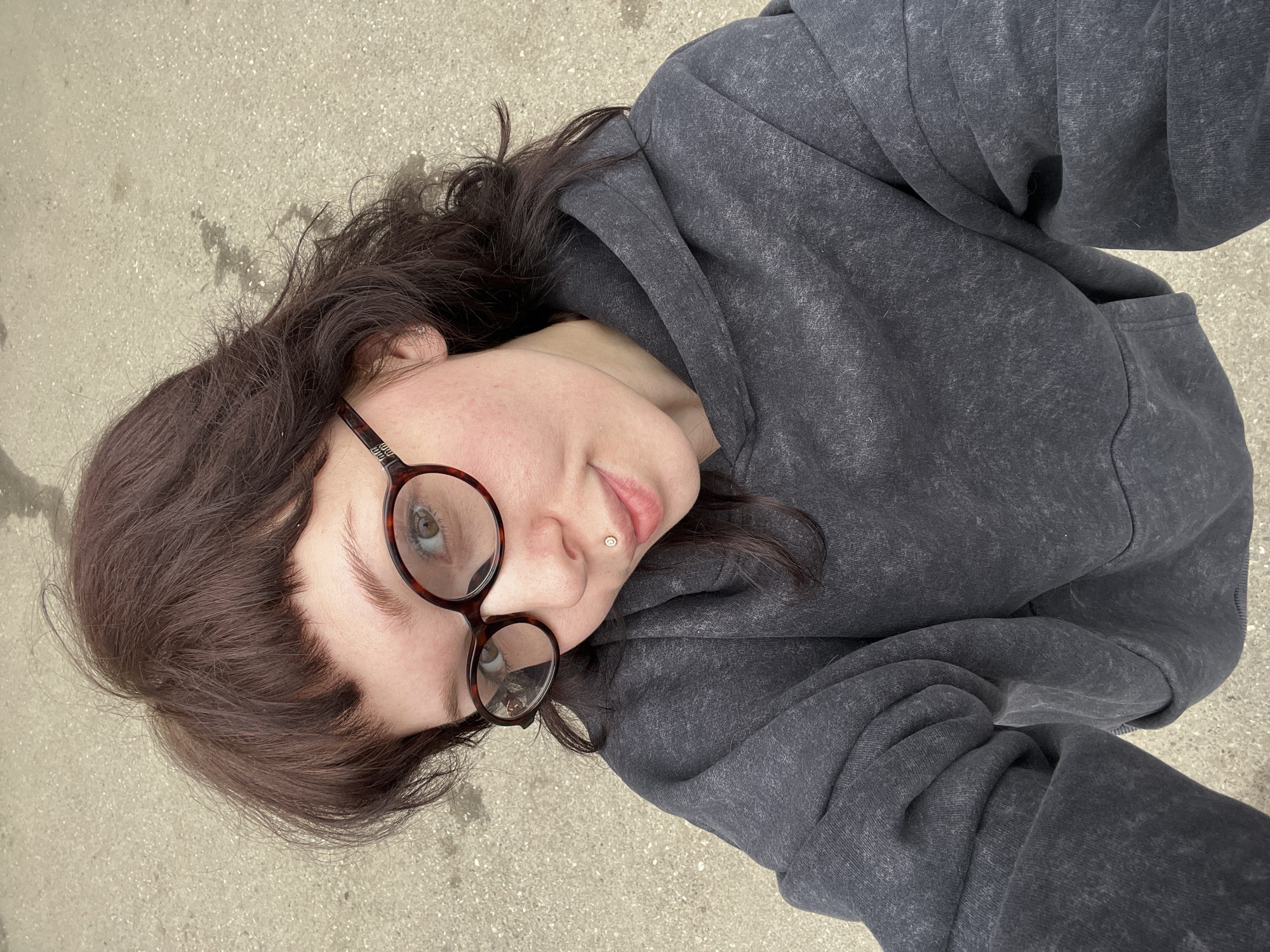 A woman with dark brown, shoulder-length hair, bangs, and wearing round, tortoiseshell-patterned glasses is the primary subject of the image. She has fair skin and a lip piercing. She is wearing a gray hoodie. The background consists of a textured gray surface, resembling concrete or pavement. The composition suggests the image was taken from a slightly elevated, angled perspective. The location details of Staraya Yum'ya, Russia, are not apparent in the image.