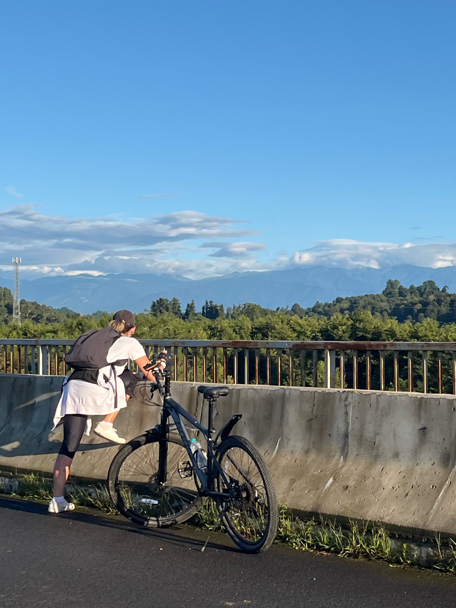 A person is bent over a bicycle on a paved road next to a concrete barrier. The person is wearing a white t-shirt, black leggings, a black cap, and a black backpack. The bicycle is black and has a water bottle attached to the frame. In the background, there are trees, mountains, and a blue sky with clouds. There is a metal railing on top of the concrete barrier. The scene appears to be outdoors, possibly during the daytime. The location is Ureki, Georgia.