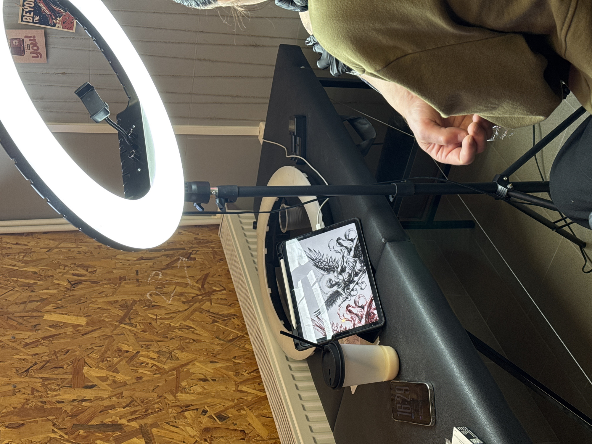A high-angle, close-up shot shows a tattoo artist preparing their equipment in a room with OSB wood paneling on one wall. A ring light is positioned to the left, illuminating the scene. A black tablet displaying a detailed drawing of a winged figure and a drink in a plastic cup with a black lid and straw are on a black padded surface. The artist, wearing a green jacket and black gloves, is visible at the top of the frame, manipulating a clear plastic wrapper or film. A black phone, showing "16:29" on the screen, is next to the cup. The precise location is Unknown, Unknown.