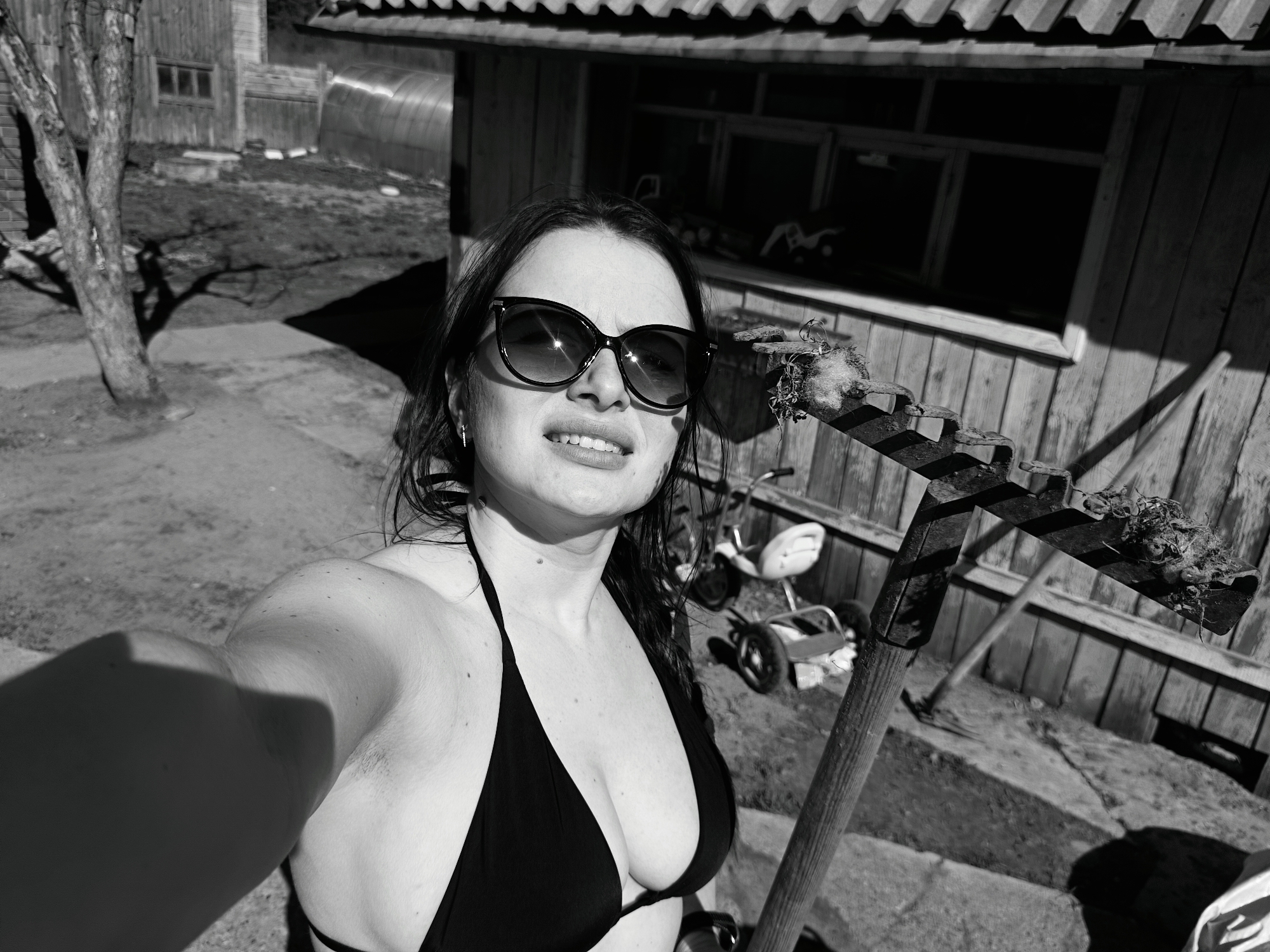 The image is a black and white selfie of a woman wearing a bikini and sunglasses. She is holding a rake. The background shows a wooden house, a yard, and some gardening tools. The location is Rusalevka, Russia.