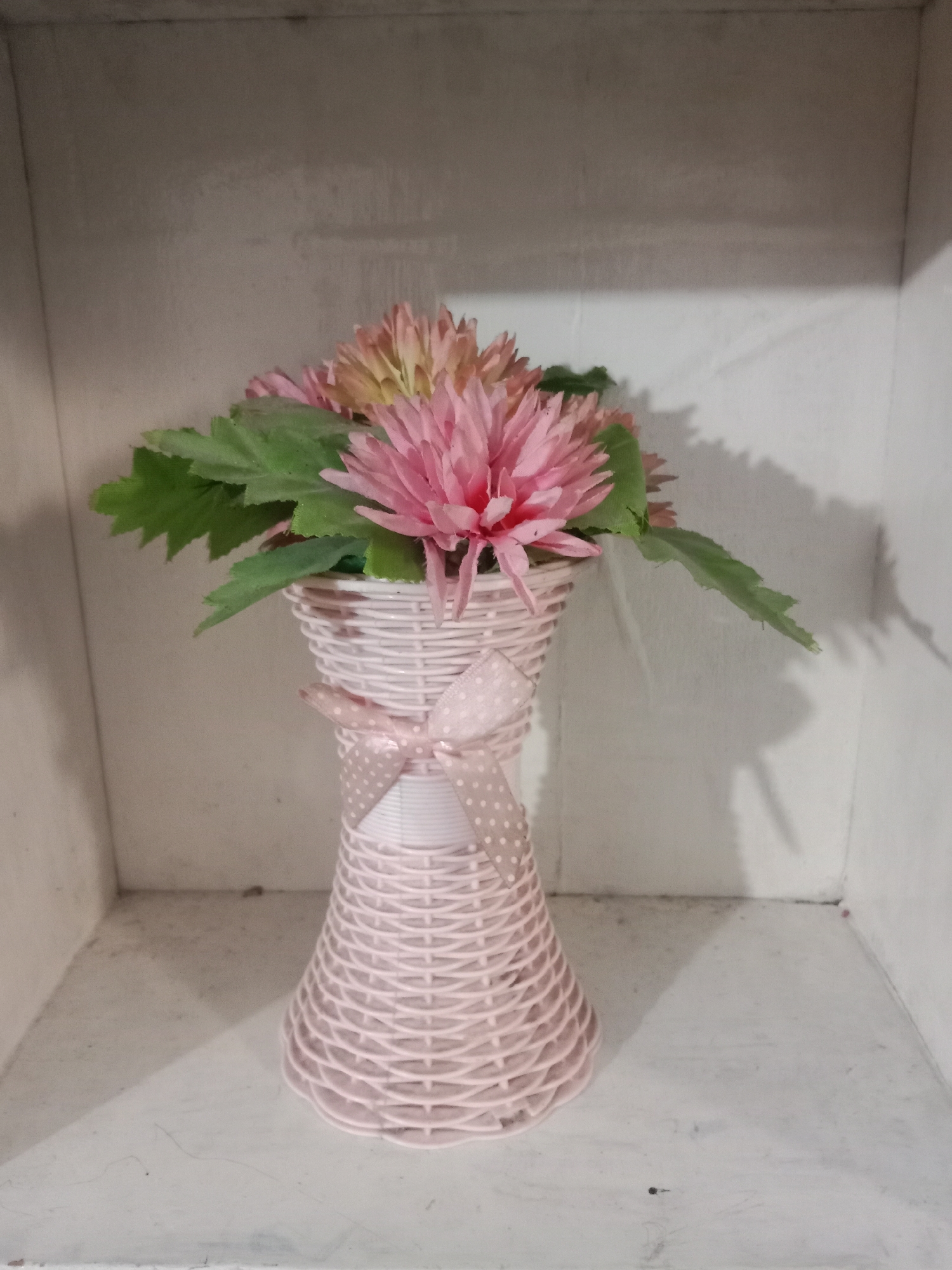 A light pink, woven vase with a polka-dot ribbon tied around its neck sits on a shelf. Inside the vase are artificial pink and yellow flowers with green leaves. The vase is positioned on a white shelf within a white shelving unit, with shadows cast on the back wall. The scene appears to be in a home or display setting, with no indication of the city of Kutosari, Indonesia.