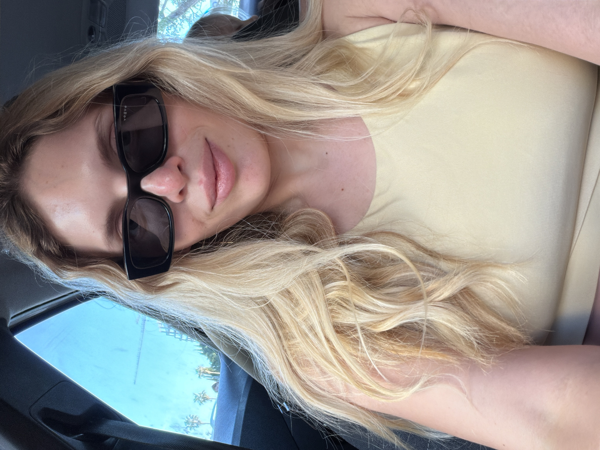 A relaxed, eye-level selfie captures a young woman with long, flowing blonde hair, likely taken by herself inside a car. Her head is tilted, and her expression is obscured by black, rectangular sunglasses. The brand "VOGUE" is subtly visible in white text on the right arm of the sunglasses.
She has full, naturally-colored lips, slightly parted. Her skin appears clear, with a few small moles visible on her shoulder and upper chest. She is wearing a simple, light yellow or cream-colored sleeveless top. Her long, wavy blonde hair cascades over her shoulder and upper body.
The setting is the interior of a vehicle, with dark-colored upholstery and elements like a seatbelt and window frame visible. Outside the car window, a bright blue sky and distant palm trees are visible, indicating sunny weather and a warm climate, consistent with the provided location context of Budva, Montenegro. The time of day is clearly daytime. The overall impression is one of a casual, stylish moment captured during a sunny outing.