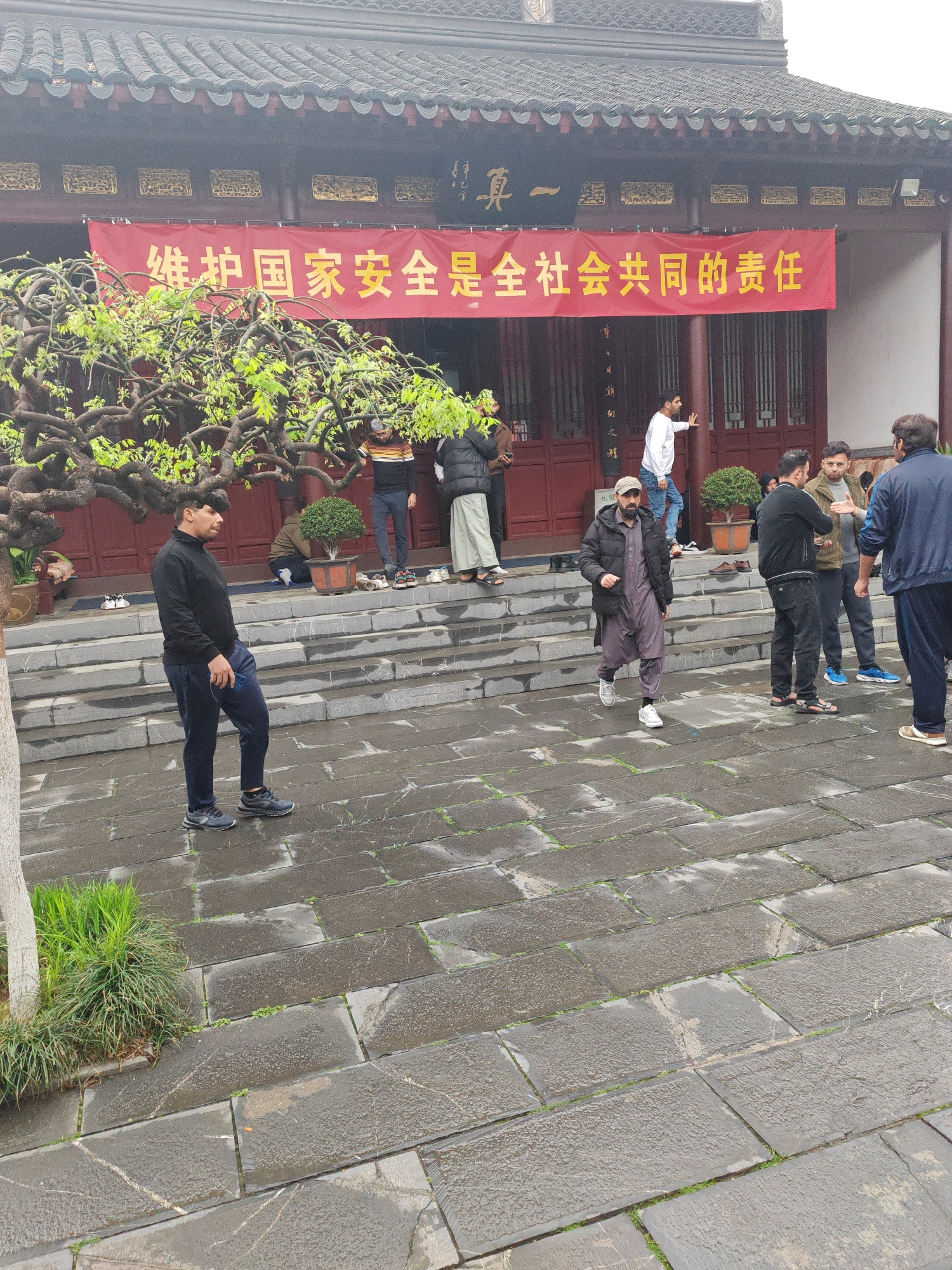 The image shows a group of people gathered outside a traditional Chinese building. A red banner with gold writing is displayed above the entrance.
People: Men are predominately featured in the image. They are of varying ages and are dressed in casual attire. Several men are standing on the steps leading to the building entrance. One man is walking in the foreground, wearing a traditional outfit and a black jacket. Another man is standing to the left, wearing black clothing and pants.
Subjects/Objects: The building has a dark roof and red columns. A tree is visible to the left of the building entrance, with branches reaching towards the roof. There are small bonsai plants in pots on the steps. Shoes are scattered on the steps outside the door.
Actions/Interactions: The men appear to be engaged in conversation and milling around. Some are standing, while others are walking or looking toward the building.
Environment: The ground is paved with large, rectangular stones that appear wet, suggesting it might have rained recently. The sky appears overcast.
Location: Based on the architectural style of the building and the cultural elements, it is likely the photograph was taken in Qinhuai District, China.