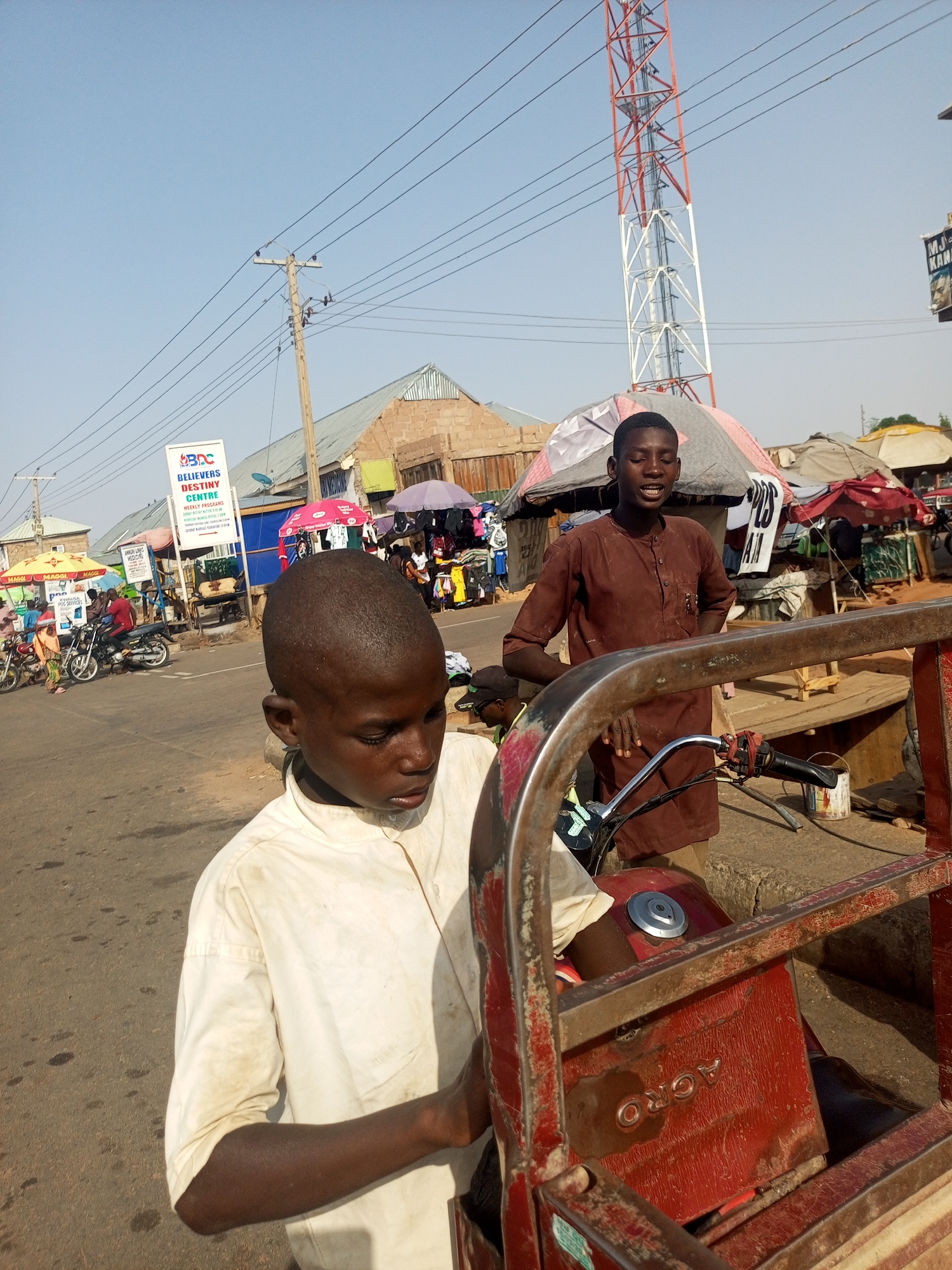 The image captures a street scene in Gombe, Nigeria, during daylight. Two young men are visible in the foreground. One is younger, wearing a soiled white shirt, and appears to be holding onto a red, rusty, three-wheeled motor vehicle with the word "ACRO" visible on its surface. The second young man stands behind the vehicle, wearing a brown shirt and looking towards the side of the image with a smiling expression.
In the background, there is a bustling street market with numerous stalls covered by various colored umbrellas. People are present, and motorcycles are parked along the road. A sign saying "BELIEVERS DESTINY CENTRE" is visible. Utility poles with numerous electrical wires crisscross the sky. A tall telecommunications tower is present, painted red and white, reaching toward the clear sky.