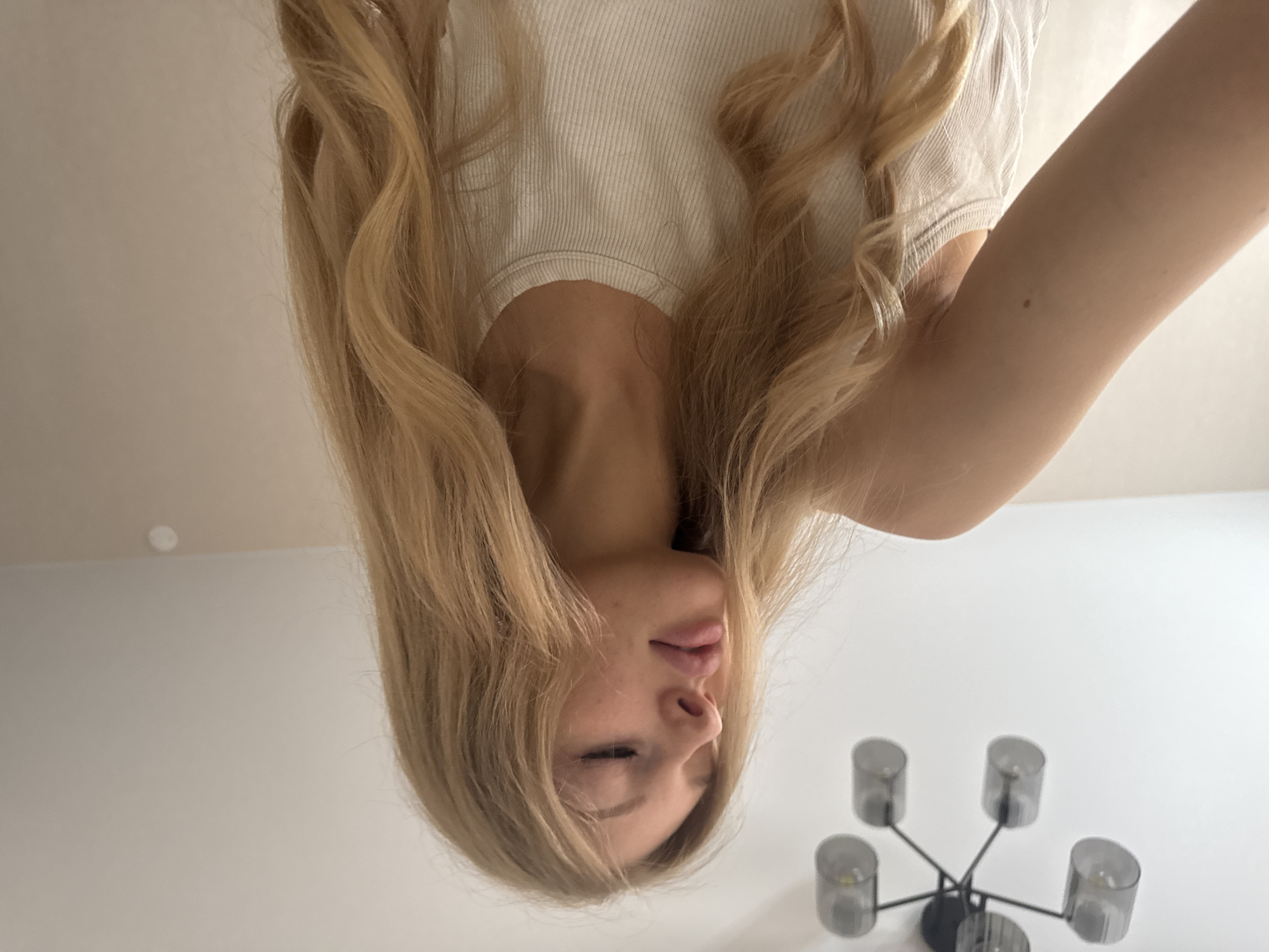 A medium shot depicts a blonde woman from an inverted perspective, likely captured with a phone or camera held above her. She is wearing a white ribbed top. Her hair is long, blonde, and slightly wavy. She appears to be lying down. The background consists of a white wall and ceiling. A decorative ceiling light fixture with multiple cylindrical shades is visible. While the woman is the primary subject, pinpointing the exact location in Perm, Russia, is not possible without additional context or visual cues from the scene.