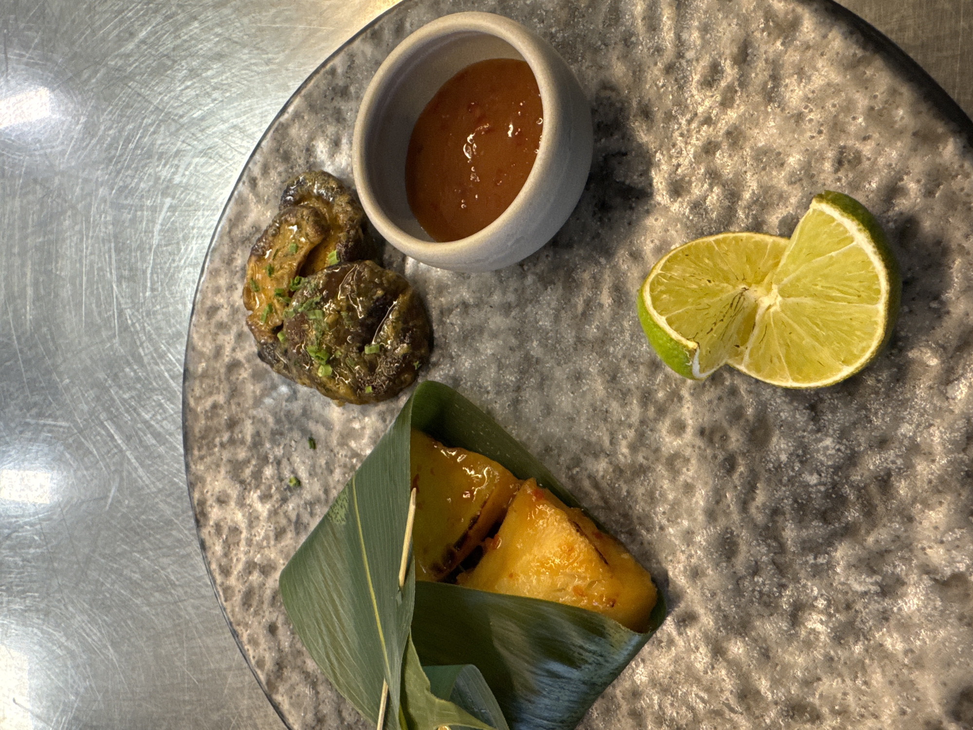 An overhead shot presents a meticulously arranged plate of food on a textured, grey, stone-like dish, set against a brushed metallic table surface. The dish features several components.
In the upper central part of the plate, a small, light grey ceramic bowl holds an amber-colored sauce, possibly a sweet chili or fruit-based condiment, with small flecks visible. To its left, a cluster of dark, irregularly shaped food items, possibly glazed mushrooms or another savory preparation, is garnished with finely chopped green herbs, likely chives. On the right side of the plate are two bright green lime wedges, cut from a single lime.
At the bottom center, a vibrant green leaf, possibly a banana or bamboo leaf, is used to wrap a portion of food. The leaf is secured with a small wooden skewer, partially revealing the contents: two pieces of a glossy, yellow-orange food item, which appears somewhat soft or sticky, possibly a type of dessert or sweet rice, glistening with a glaze.
The presentation suggests a refined culinary experience, possibly Asian-inspired or fusion cuisine, suitable for a restaurant setting in Sofia, Bulgaria. The lighting appears artificial, highlighting the textures of the food and plate. No text is visible in the image.