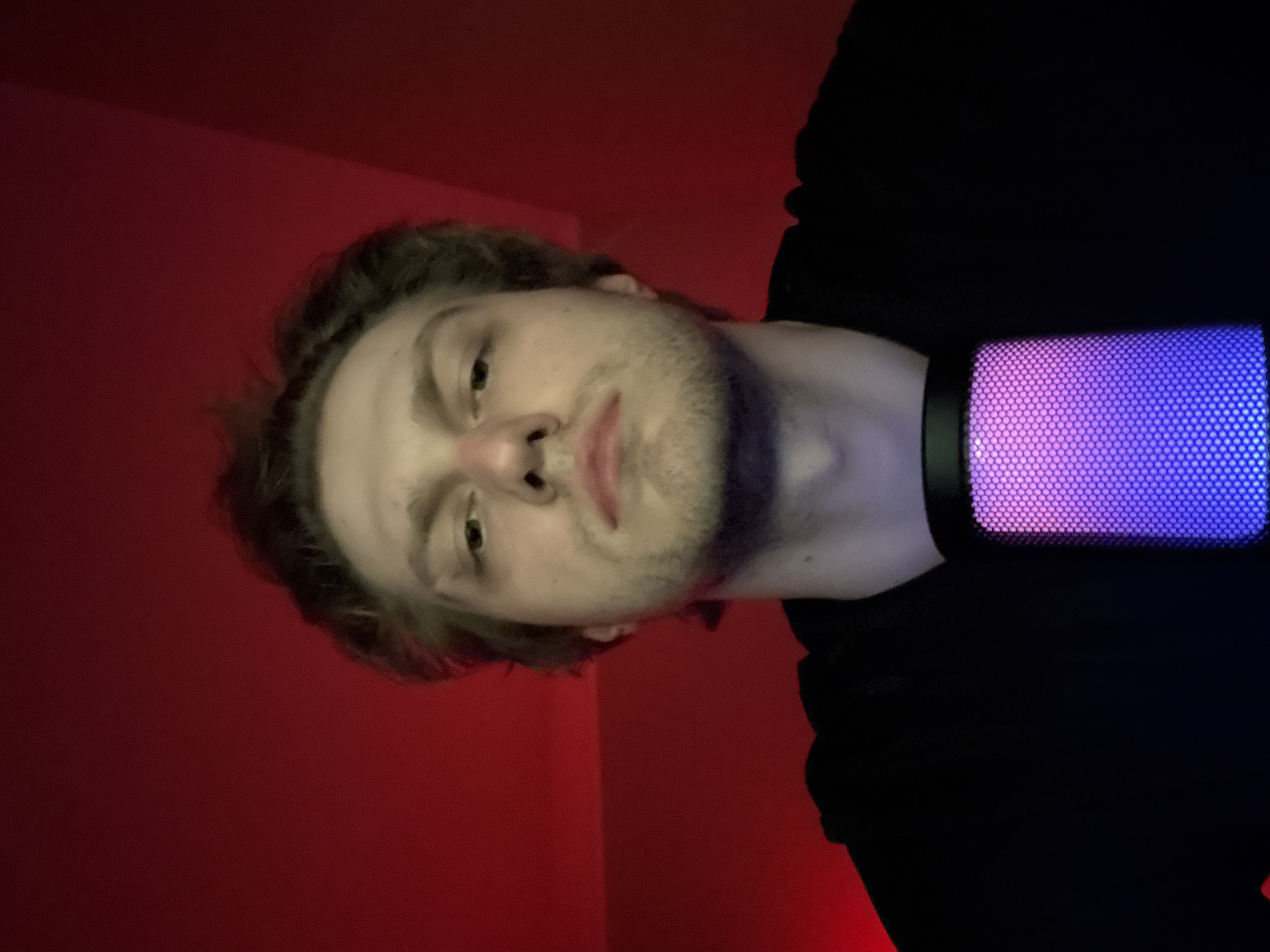 A medium shot portrait of a young adult male with light brown hair and a short beard is captured against a red background. He is wearing a black t-shirt and looking directly at the camera with a neutral expression. In the foreground, a microphone with purple and pink LED lighting is positioned in front of him, partially obscuring his lower face and neck. The lighting in the image is predominantly red, with the microphone emitting a vibrant glow. The setting appears to be indoors, possibly a studio or home environment, with the red background suggesting ambient lighting or a deliberate backdrop. There is no information to suggest the location is in Khimki, Russia.