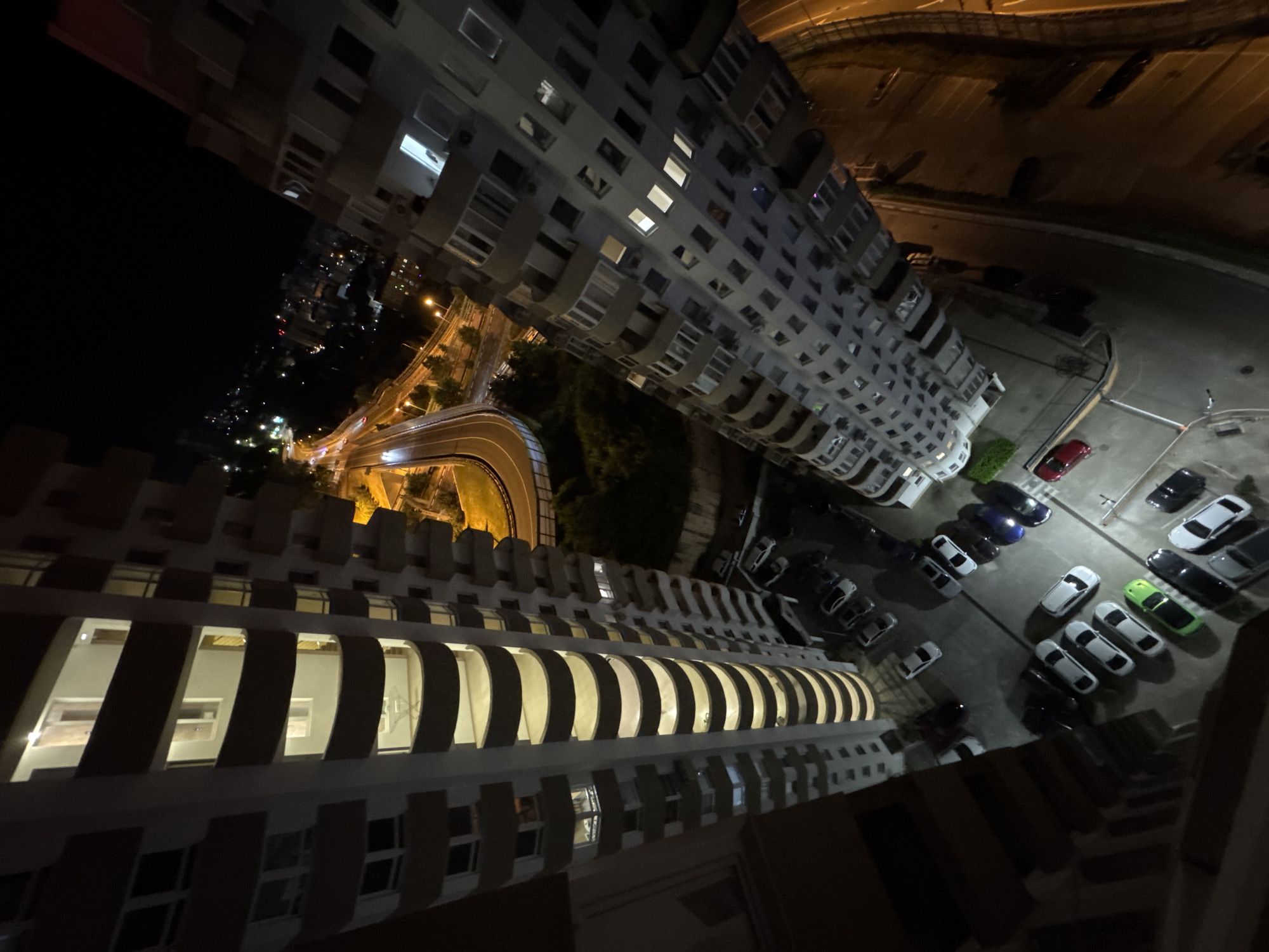 This image depicts a nighttime aerial view of a residential area in Sochi, Russia. Two large, modern apartment buildings dominate the foreground, their balconies and windows illuminated by interior lights. One building, on the left, is characterized by its curved balconies and a yellowish glow emanating from within. The building on the right has a more angular facade with numerous windows, many of which are also lit.
Below the buildings, a parking lot is visible, containing several cars of various colors, including white, black, silver, and a distinctive bright green car. The parking lot appears to be paved. Adjacent to the buildings and parking lot, a curving roadway with illuminated streetlights is prominent, suggesting a city highway or major thoroughfare. Beyond the immediate vicinity of the buildings, the distant cityscape of Sochi unfolds, with scattered lights indicating further urban development stretching towards the horizon. The dark night sky serves as the backdrop, with no celestial bodies clearly visible. The overall impression is one of a well-developed, illuminated urban environment at night.