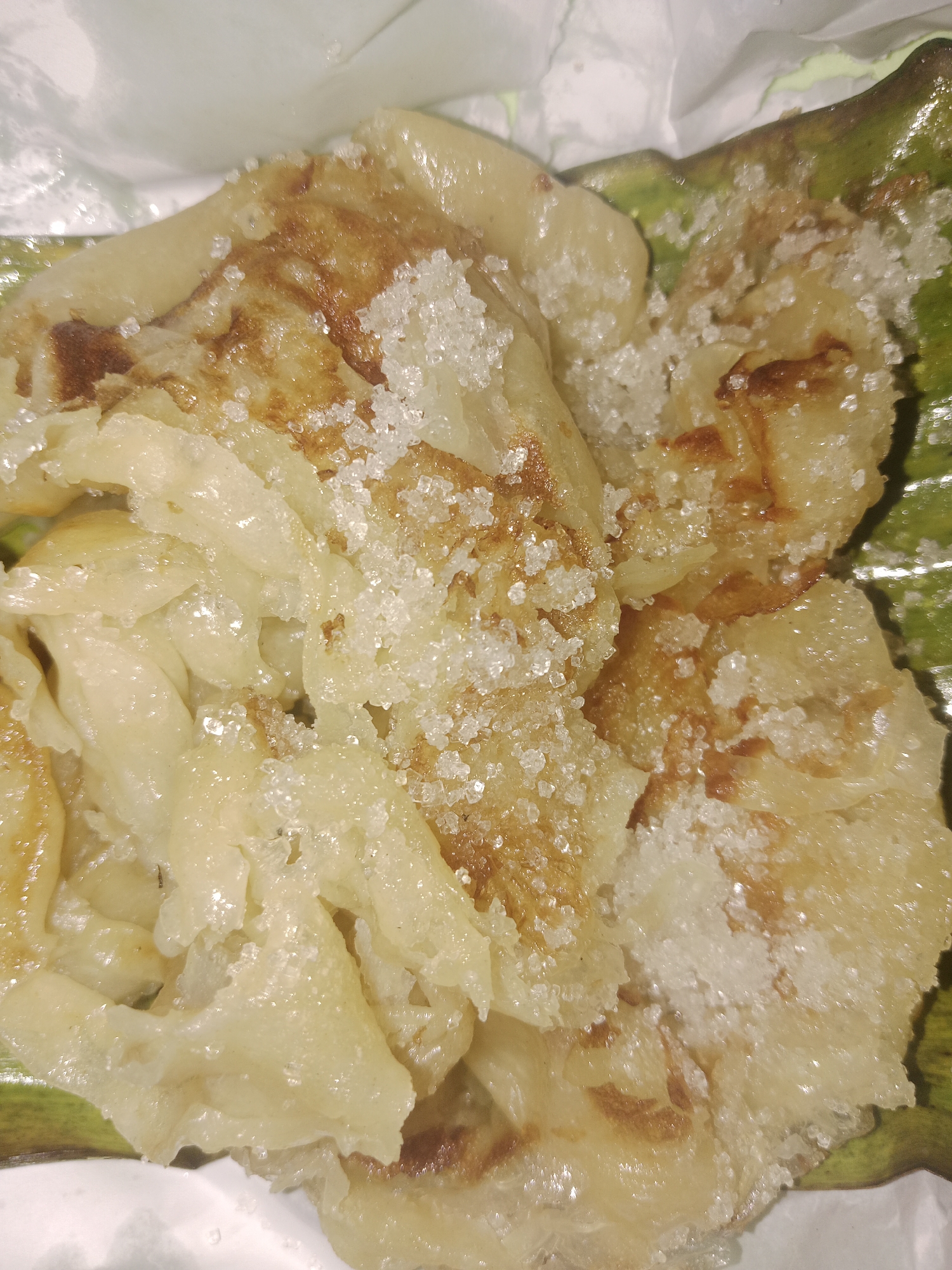 A close-up, overhead shot captures a portion of what appears to be a sweet, flaky pastry, possibly a type of pancake or crepe, sprinkled with coarse sugar crystals. The pastry exhibits a golden-brown, slightly caramelized exterior, indicating it has been cooked. It is resting on a green, textured surface, possibly a banana leaf, which is itself on a white, possibly paper, wrapping. The background is indistinct and light-colored. The overall impression is that of a close-up food photograph, likely taken in a domestic or street-food setting in Meunasahara, Indonesia, showcasing a local delicacy.