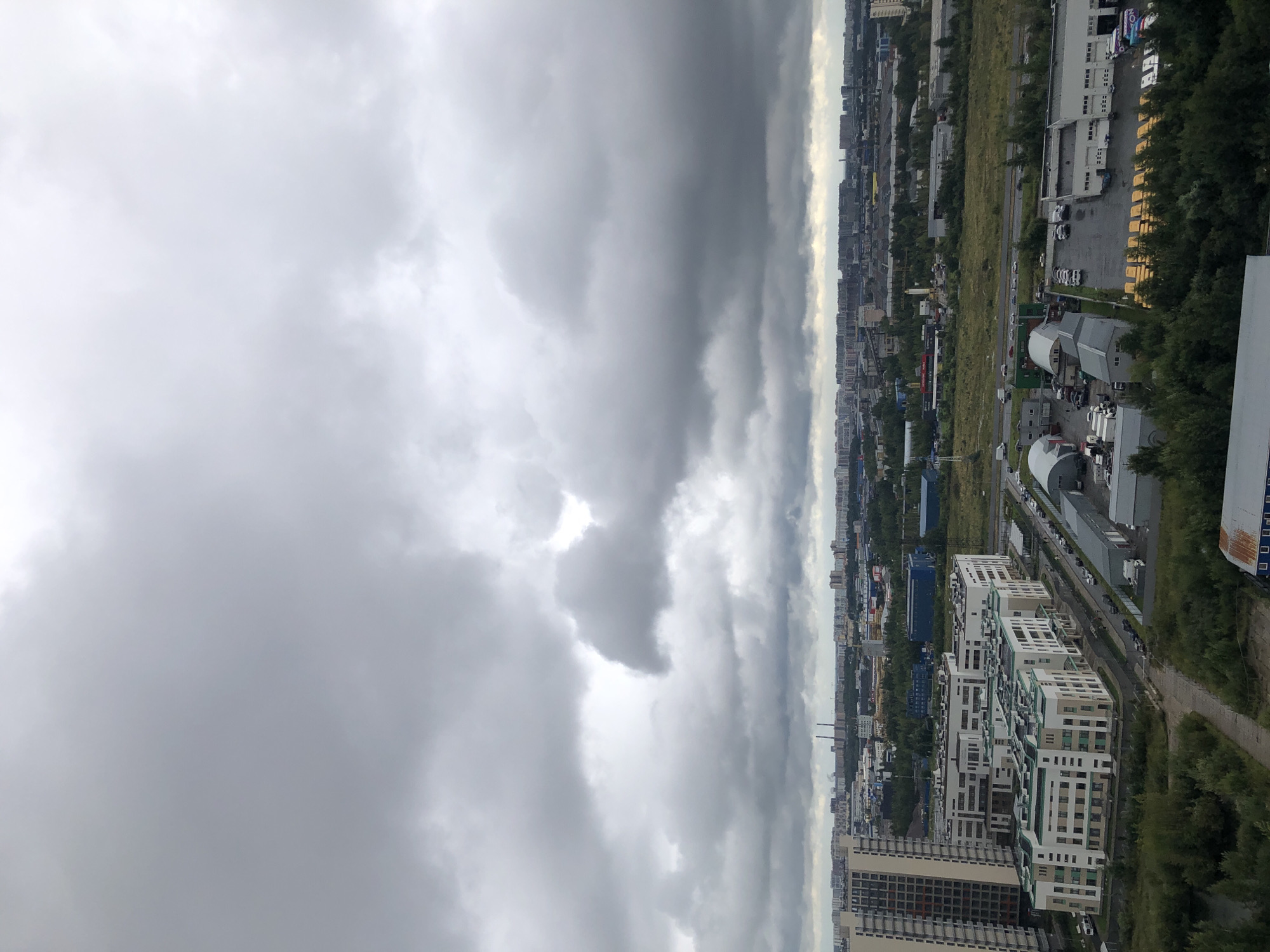 This is an aerial photograph of an urban landscape under a cloudy sky. The foreground features a cluster of modern, light-colored residential buildings with green accents on their facades. Adjacent to these buildings are several industrial structures, including warehouses with grey roofs and white siding, some with loading bays. Parking lots surrounding these structures are filled with various vehicles, including cars, vans, and trucks. A multi-lane road is visible, with cars driving on it. In the midground and background, more buildings, including taller residential towers and industrial complexes, stretch across the horizon. The cityscape is interspersed with patches of green vegetation, including trees and grassy areas. The sky is dominated by dense, grey clouds, suggesting overcast or potentially rainy weather. The overall scene depicts a mixed-use urban environment with residential and industrial zones under a somber sky.