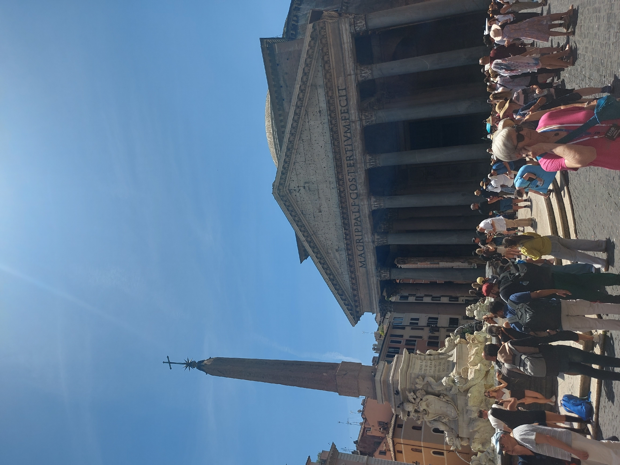 The image depicts a daytime view of the Pantheon in Rome, Italy, and the Piazza della Rotonda. The Pantheon's facade, including its columns and inscription "MAGRIPPALF-COS TERTIVM FECIT," is visible. A crowd of tourists stands near the entrance of the Pantheon. To the left of the Pantheon, the Obelisco Agonale rises into the blue sky. A fountain adorned with sculptures stands between the obelisk and the Pantheon. The inscription "CLEMENS XI PONT-MAXONTIS ET FORGINAMENTO ANND SAL MDCCXI PONTIF-X" can be seen on the fountain.
