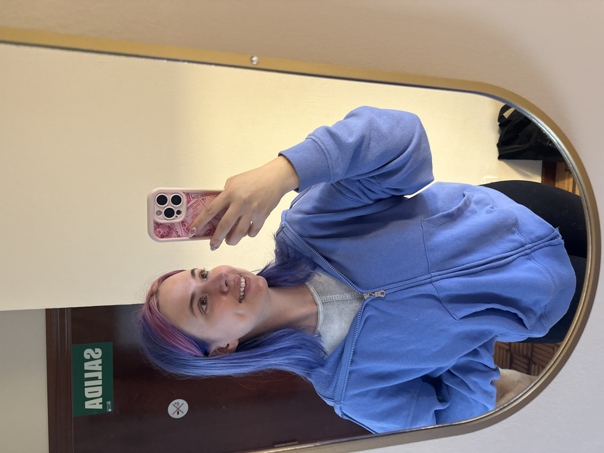 A young woman takes a selfie in a mirror. The reflection shows her facing the mirror with her right side slightly more prominent. She is holding a pink smartphone. The woman has shoulder-length blue and pink hair. She wears a light blue zip-up hoodie over a gray shirt. The background includes a door with the word "SALIDA" in green lettering. There is also a no smoking symbol. The location is possibly in Gijón/Xixón, Spain, given the "SALIDA" sign.