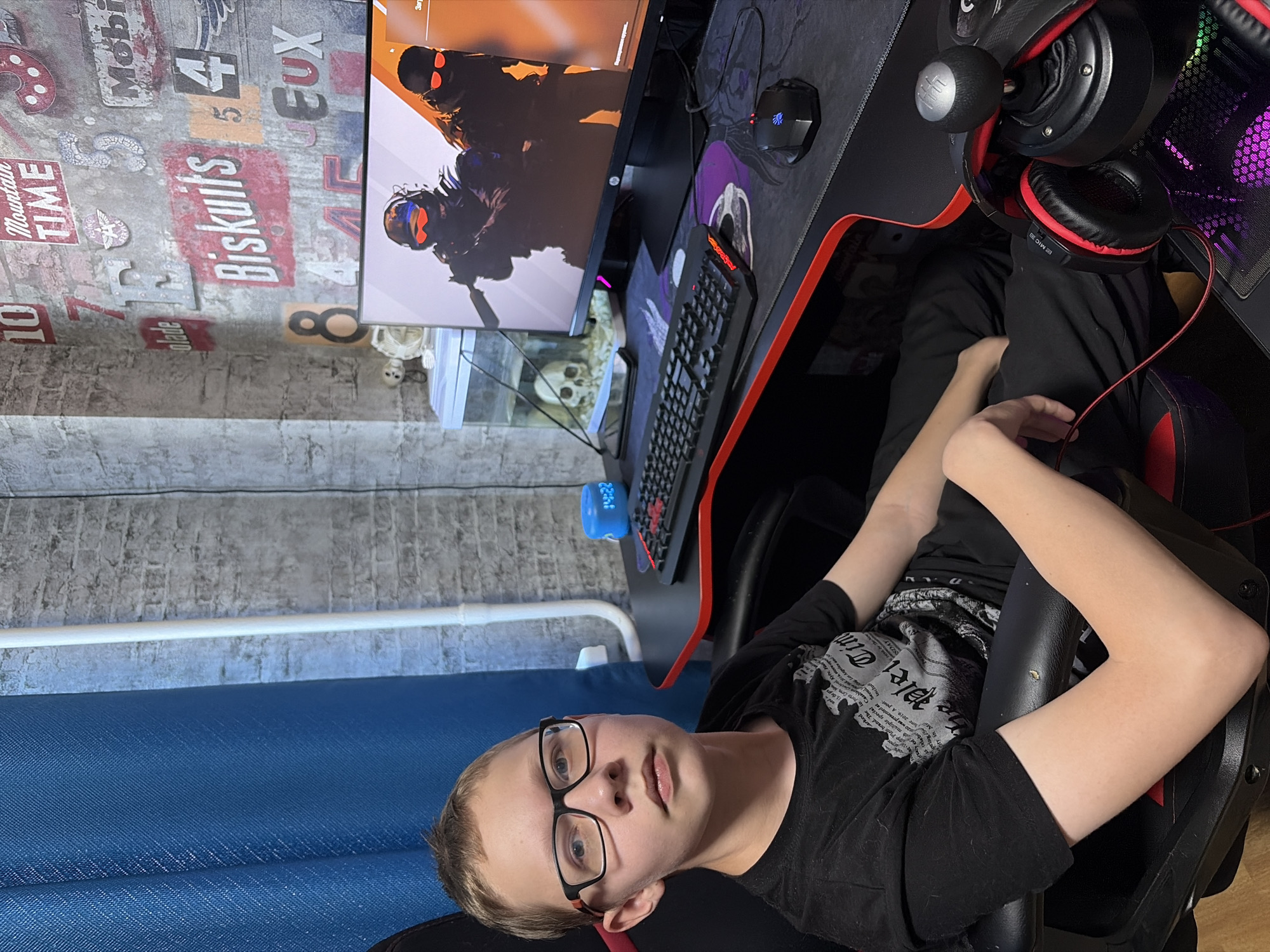 A young person wearing glasses and a black t-shirt sits in a gaming chair in front of a gaming setup. The setup includes a computer monitor displaying a video game, a keyboard, a mouse, a steering wheel controller, and headphones. A small skull decoration is visible under the monitor. The background features a wall with decorative lettering and a blue curtain. The location is Chaikovsky, Russia.