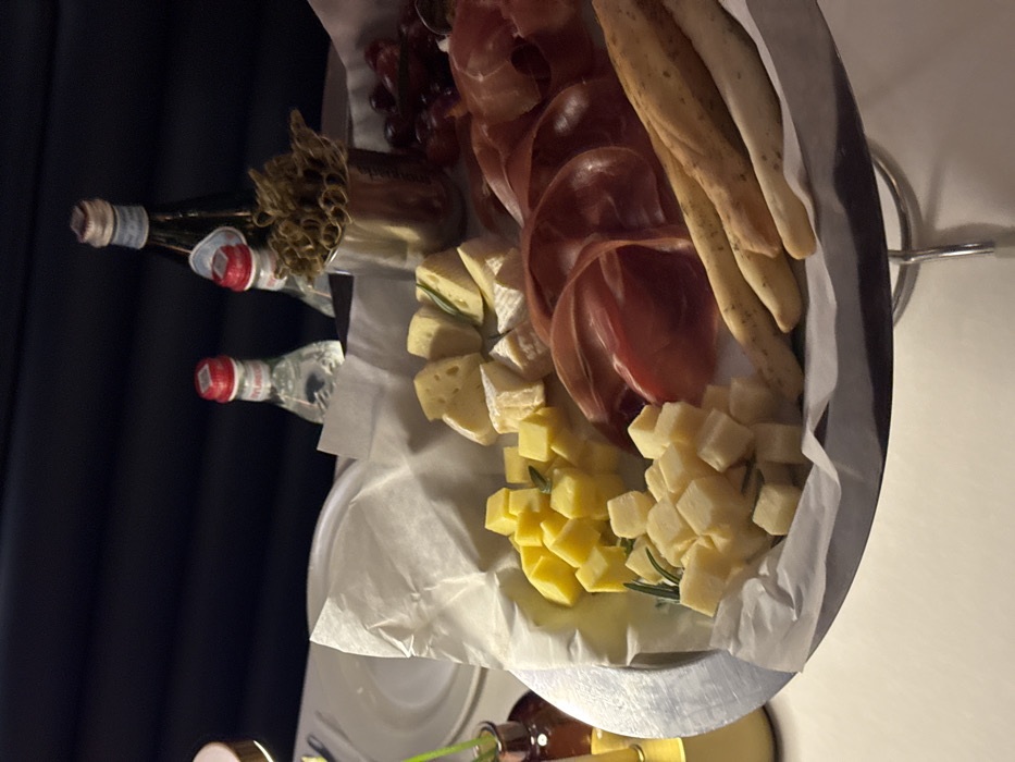 The scene depicts a charcuterie and cheese platter presented on an elevated, circular, silver-colored stand atop a white tablecloth in an indoor setting within a city in Yerevan, Armenia. The platter is lined with crumpled white paper and holds multiple food items. These include a collection of uniformly cubed yellow cheese, a pile of uniformly cubed light yellow cheese, and several wedge-shaped pieces of soft, white-rinded cheese (resembling brie), some garnished with small green rosemary sprigs. Adjacent to the cheeses are multiple slices of reddish-brown cured meat, folded and layered. Several long, thin breadsticks (grissini) are arranged along the edge of the platter. To the upper right of the platter, a small cluster of red grapes is visible.
In the background, positioned behind the platter, are three bottles and a metallic cup. One tall, dark green bottle with a light blue label is identifiable as San Pellegrino sparkling water. To its left, two smaller clear bottles with red caps and light green labels are visible, identifiable as Ayruni mineral water. A small, cylindrical, metallic container holds numerous thin, curly, dried brown botanical elements. To the left foreground, a small, dark amber glass vase containing green foliage is partially visible, next to a white ceramic dinner plate. The immediate background consists of a dark, horizontally paneled wall, suggesting a commercial establishment such as a restaurant or bar. The lighting is dim, casting soft illumination on the foreground objects. No individuals are present or visible in the image.