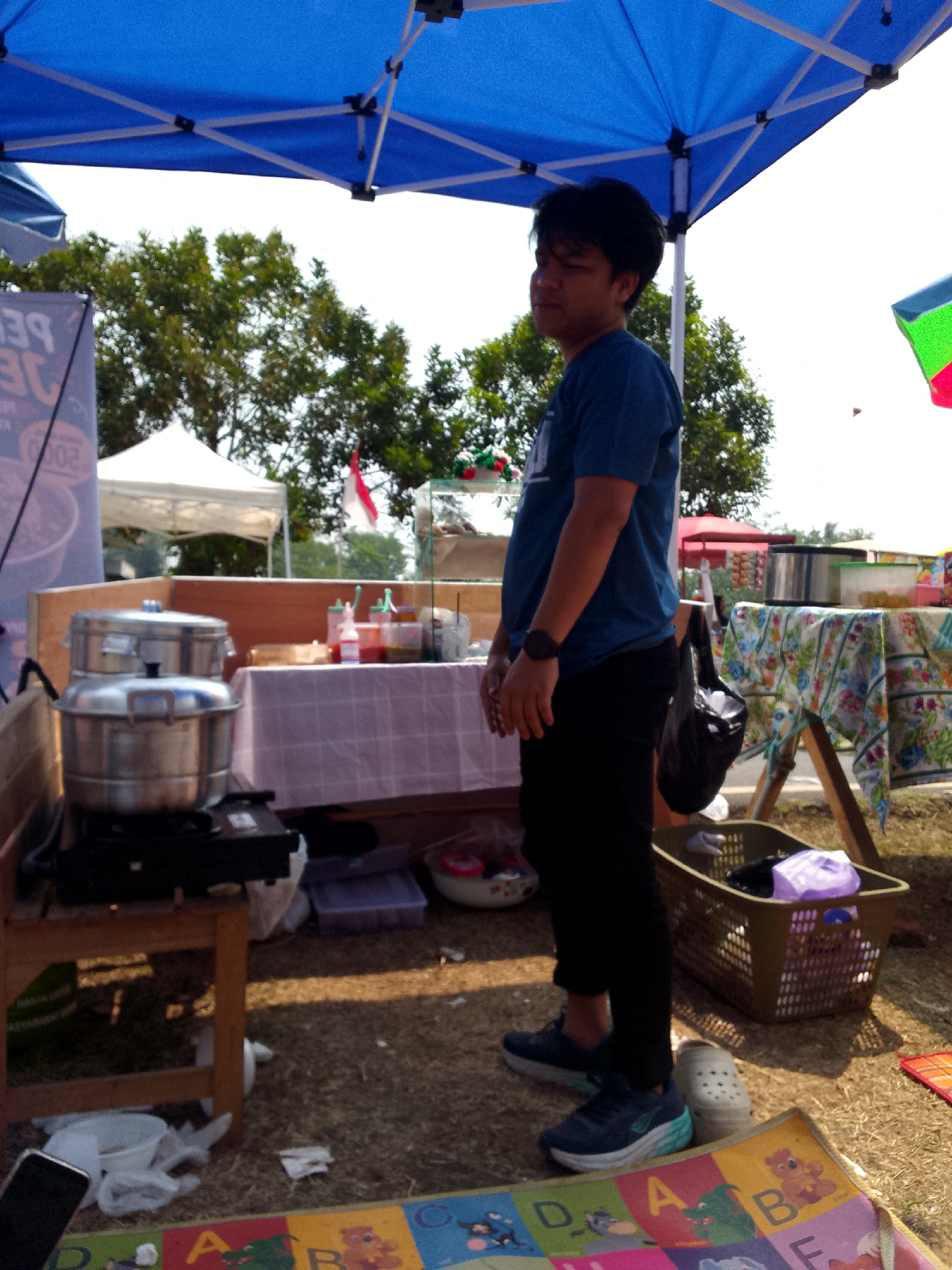 A young man in a blue t-shirt and black pants stands in front of a food stall under a blue canopy. He is wearing a watch on his left wrist and blue and white sneakers. To his left, a two-tiered stainless steel steamer sits on a wooden stand. Behind the steamer, a table is covered with a pink and white checkered cloth, and several bottles of condiments are visible. In the background, trees and two white tents are set up. To the man's right, a colorful blanket with letters and cartoon characters is spread on the ground. A woven basket filled with items, including a purple bag, sits beside the blanket. The scene appears to be an outdoor market in Mandalawangi, Indonesia, with a bright, sunny sky.