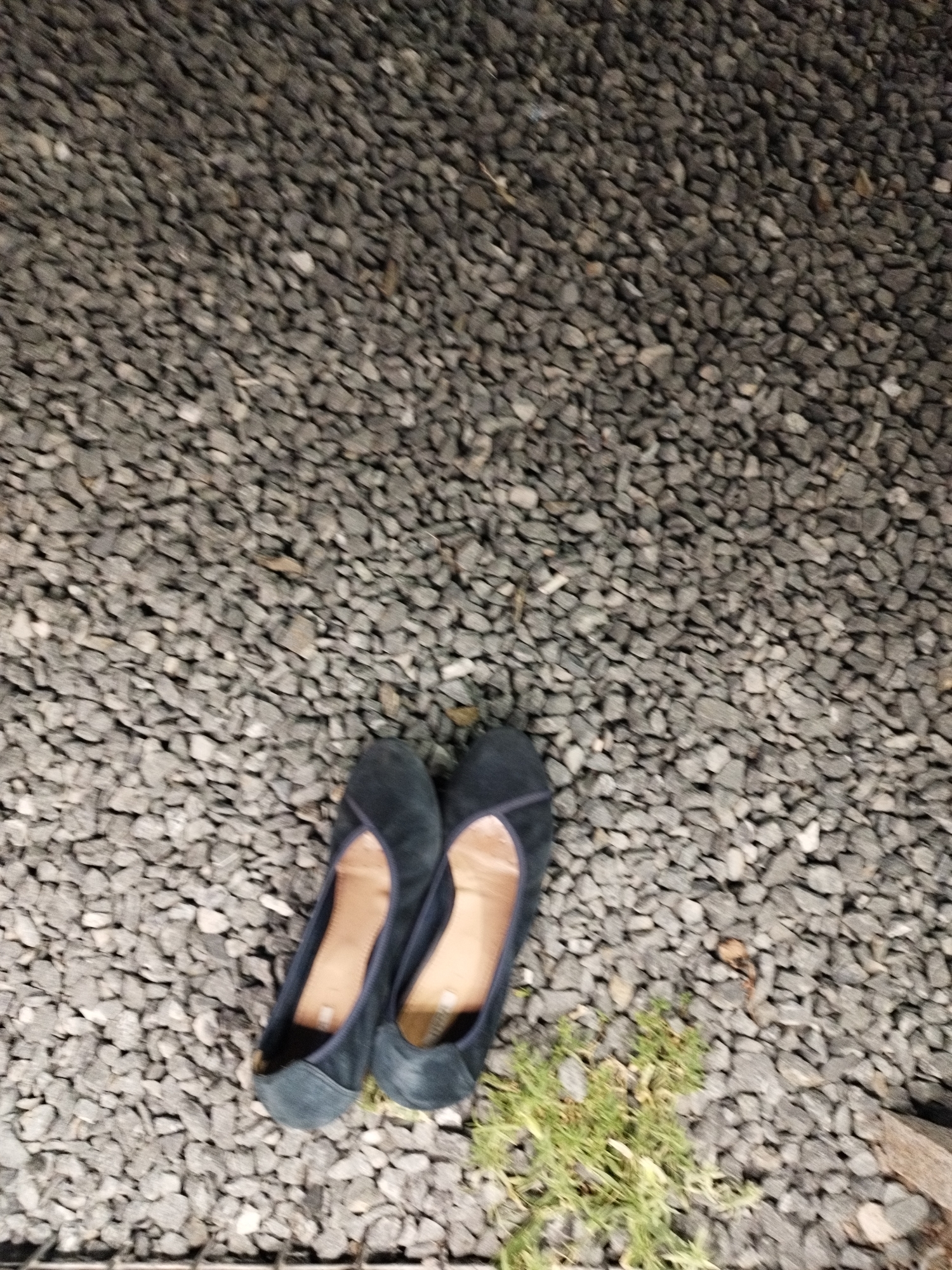 A pair of dark blue ballet flats are placed on a surface of grey gravel. The shoes are positioned side-by-side, with the right shoe slightly overlapping the left. The gravel surface is rough and uneven, with small stones of various shades of grey. A small patch of green grass is visible near the shoes. The lighting suggests it is daytime, but the scene is not brightly lit. The background is entirely composed of the gravel. There is no indication of the location being in Astana, Kazakhstan.