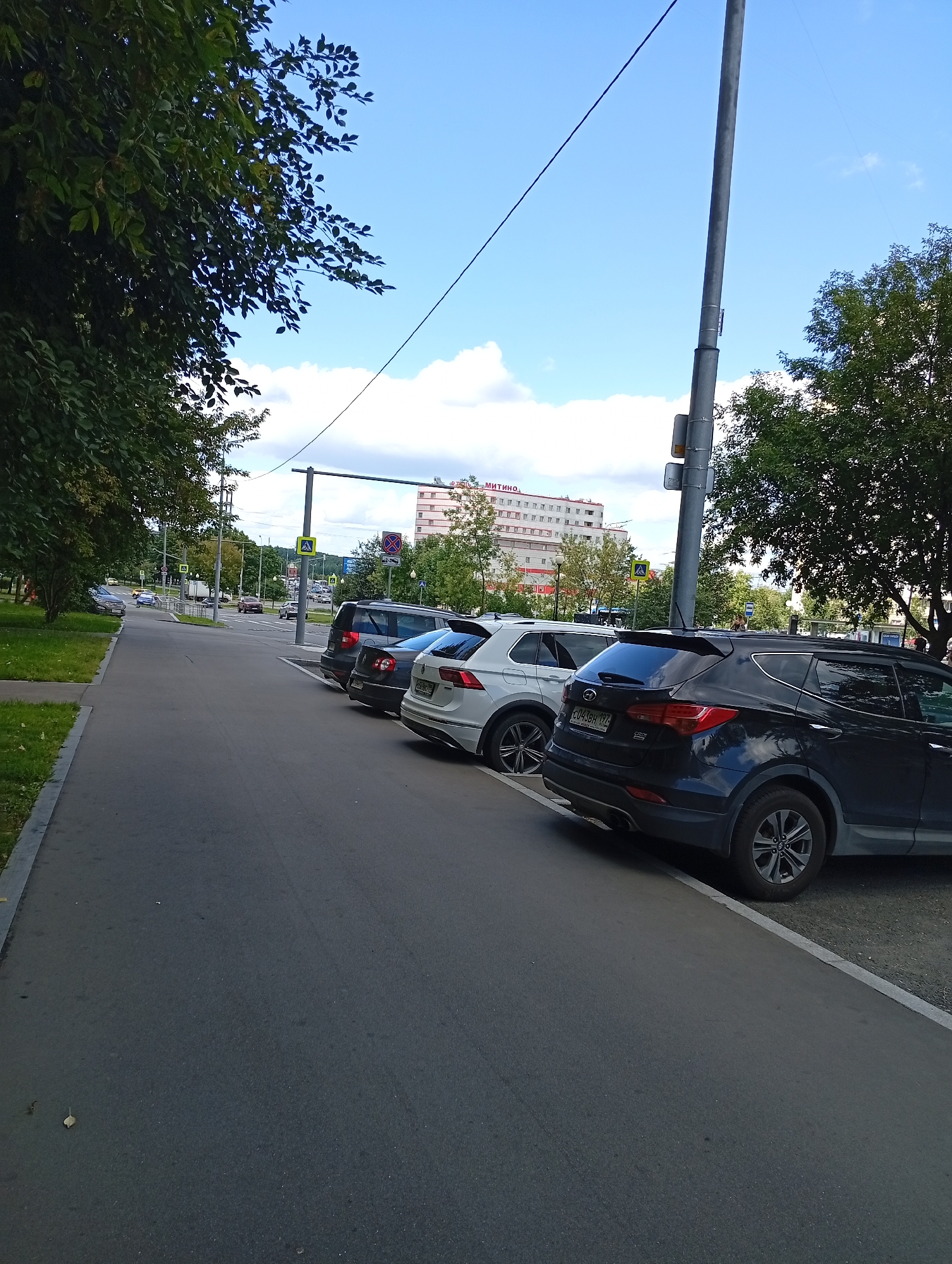 A daytime, eye-level outdoor shot captures a street scene in an urban environment, likely on a clear day with scattered clouds. In the foreground, a dark blue SUV is parked alongside a paved road, its rear end and wheels visible. To its left, a white SUV is also parked, followed by another dark vehicle. The road curves to the left, revealing a pedestrian crossing with a yellow diamond-shaped sign depicting a person walking. Further down the road, cars and a white truck are in motion. On the left side of the road, a grassy area is bordered by a sidewalk and a line of trees. In the background, a multi-story building with visible windows and a sign that appears to read "МИТИНО" stands under a bright blue sky. A tall metal pole with what looks like a traffic light or signal box is positioned near the parked vehicles. The scene conveys a sense of everyday urban life.