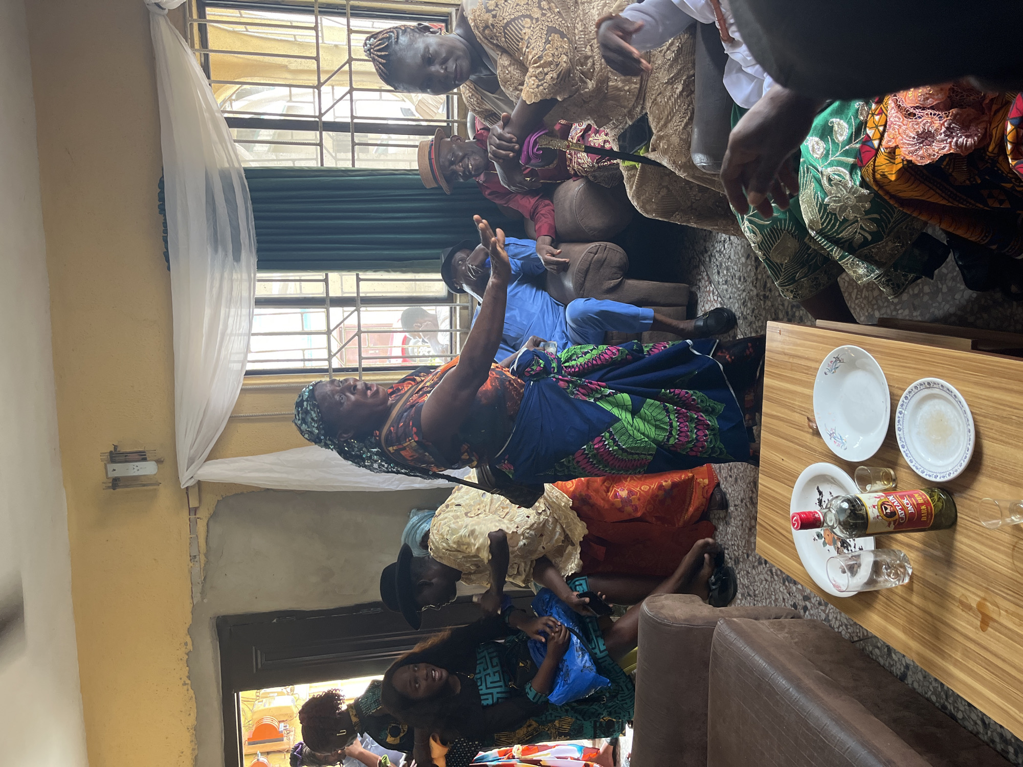 Location: Warri, Nigeria.
The scene depicts a gathering indoors, likely a celebration or family event. Several individuals are present, dressed in traditional Nigerian attire featuring vibrant patterns and colors. One woman stands and gestures with her hands raised, possibly addressing the group. Others are seated, observing the speaker. A table holds plates, glasses, and a bottle of liquor, suggesting food and drinks are part of the gathering. The room appears to be simply furnished with curtains covering the windows.