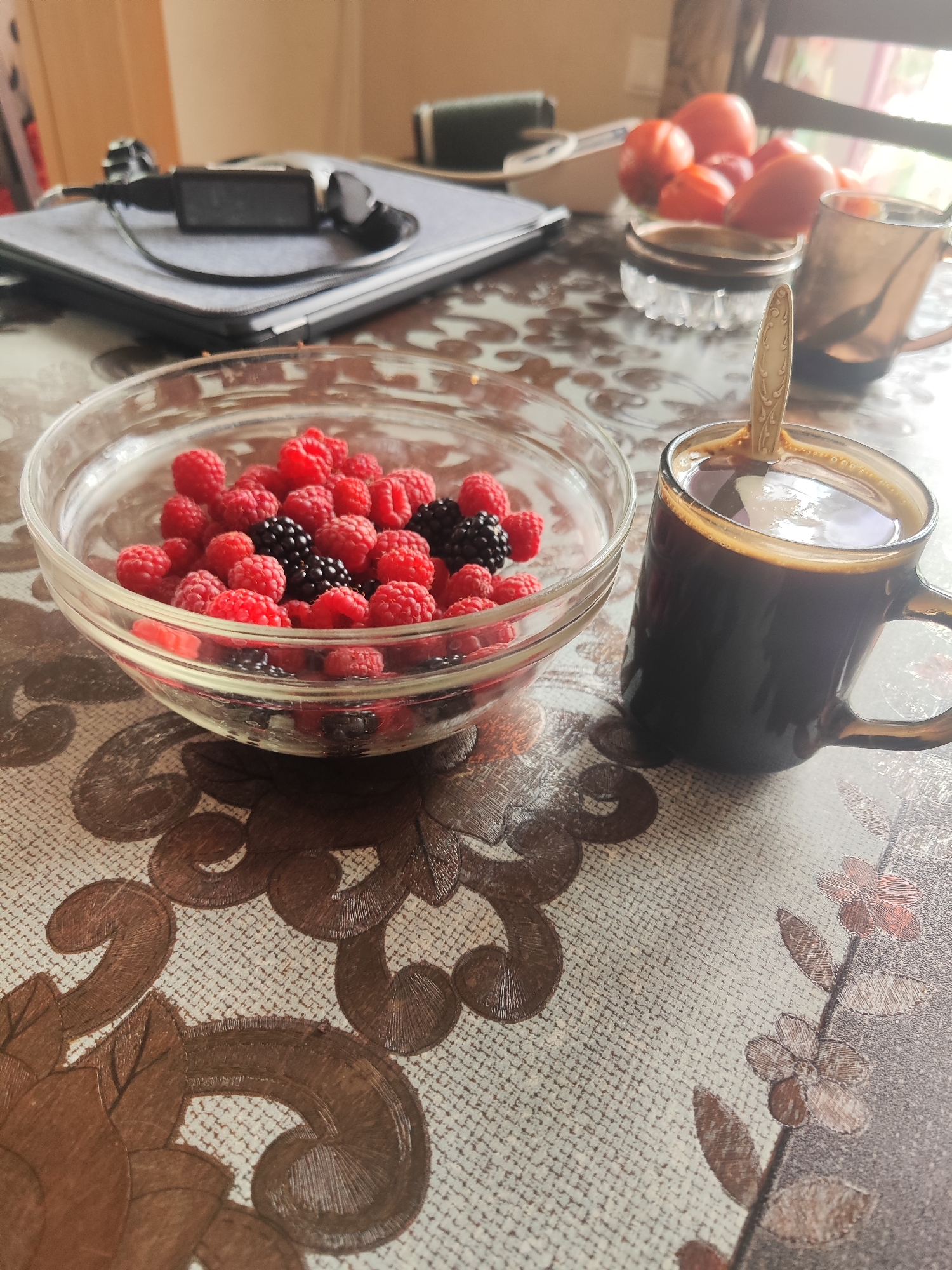 A bowl of raspberries and blackberries sits on a patterned tablecloth next to a mug of black coffee with a spoon. In the background, a tablet with a smartwatch on it rests on a surface, along with some fruit and another mug. The scene appears to be indoors, on a table. The location is Gvardeysk, Russia.