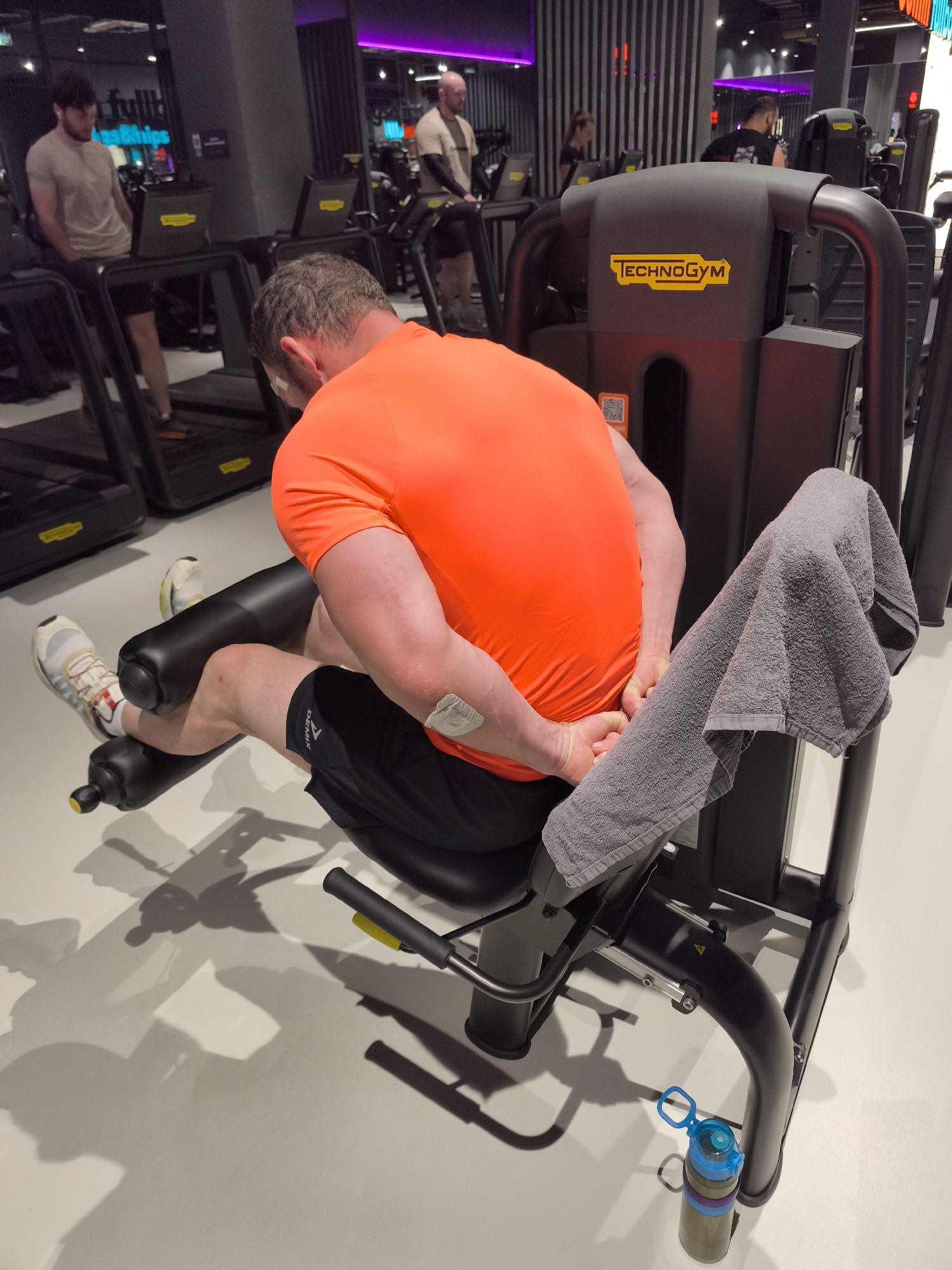 The image shows a person in a gym setting, using a Technogym leg extension machine. The person is wearing an orange t-shirt, black shorts, and athletic shoes. A gray towel is draped over the machine. A water bottle is on the floor next to the machine. In the background, other gym equipment, including treadmills, is visible, with other people exercising. The location cannot be ascertained from the image, and there is no indication that the image was taken in Moscow, Russia.