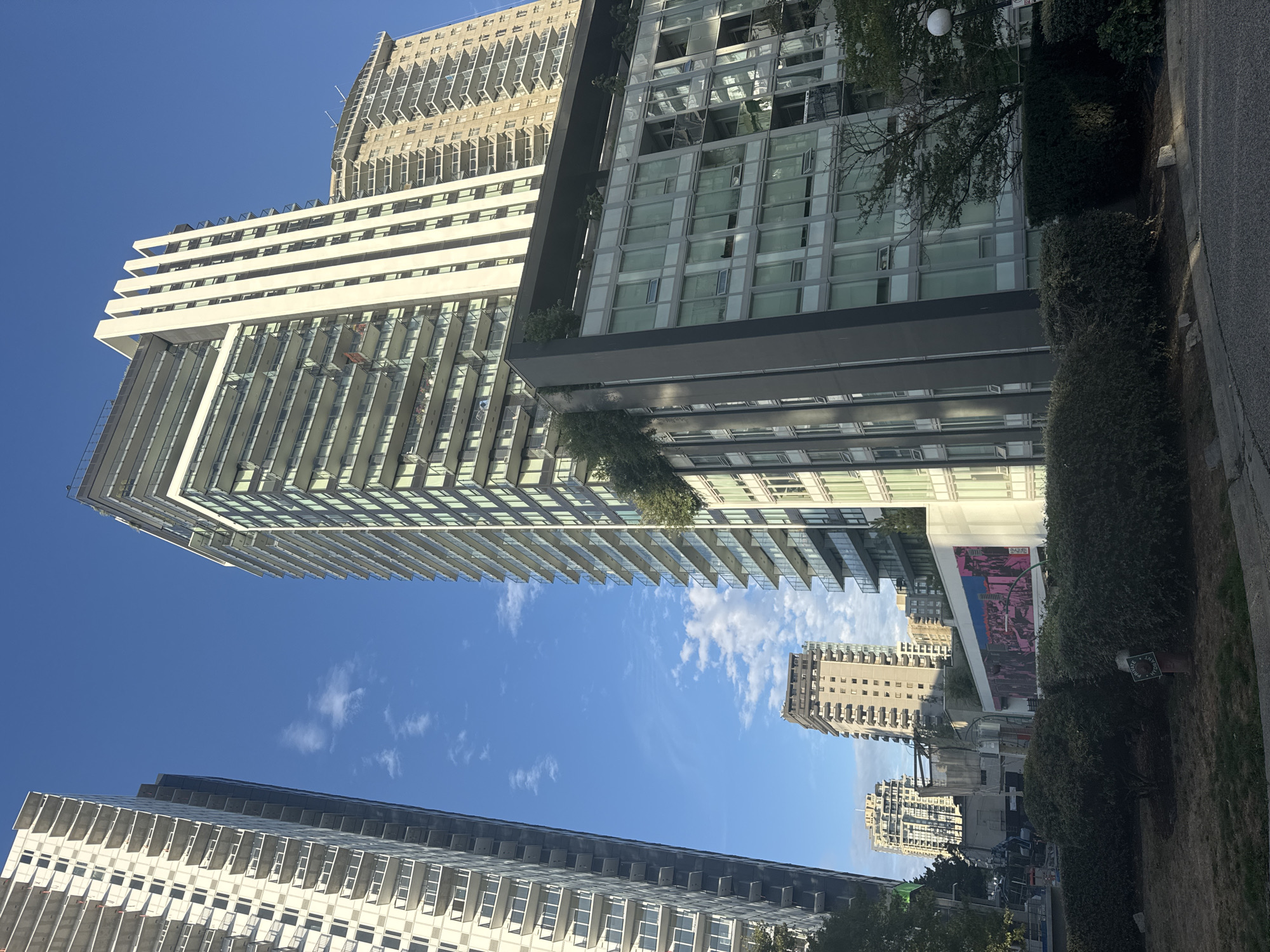 A low-angle, wide shot captures several modern high-rise buildings under a bright blue sky with scattered white clouds. The buildings are a mix of residential and possibly commercial spaces, characterized by their glass facades, balconies, and concrete structures. In the foreground, a manicured hedge runs across the lower portion of the frame, with a fire hydrant visible on the left side. The scene appears to be in an urban setting, likely a city center, with a focus on contemporary architecture.
This image was taken in the city of New Westminster, Canada. Specifically, it is believed to be in the downtown core, near the waterfront, possibly along Columbia Street or Agnes Street, where a number of new developments have been erected in recent years.