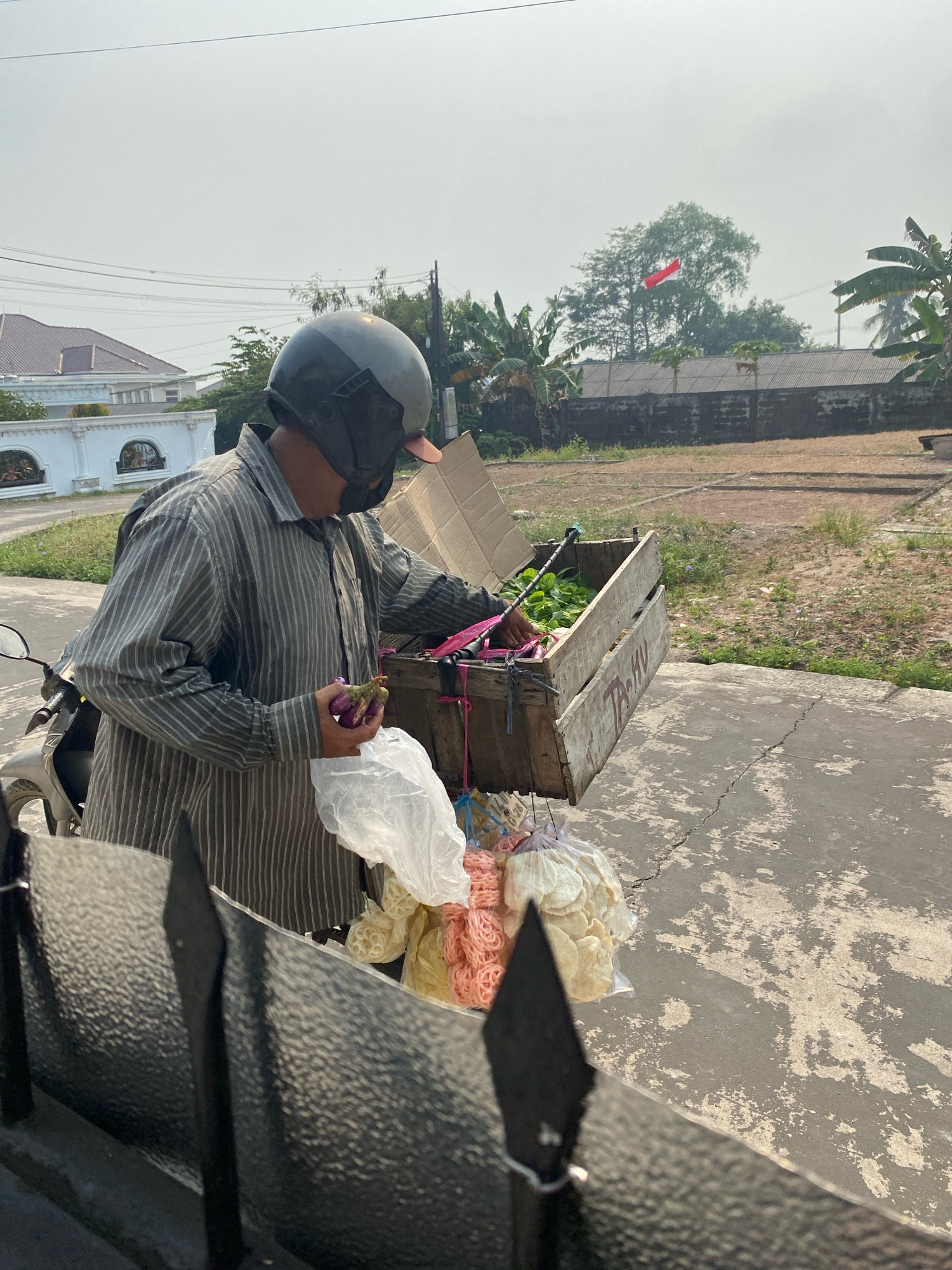 A person wearing a striped long-sleeved shirt and a helmet is standing next to a motorcycle, holding two eggplants and a plastic bag. They are reaching into a wooden crate filled with vegetables. The crate is attached to the back of the motorcycle and has pink ropes securing some of the contents. In front of the crate, several bags of snacks, possibly crackers, are hanging. The scene appears to be a street in Palembang, Indonesia, with a house and trees in the background. The sky is hazy, and the ground is paved with concrete. A red and white Indonesian flag is visible on a pole in the distance. The overall setting suggests a vendor preparing to sell produce.