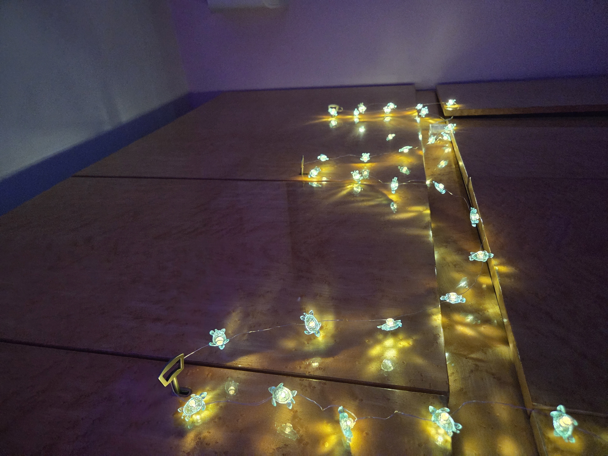 The image shows a string of turtle-shaped fairy lights draped across a wooden surface, likely a headboard or cabinet. The lights are illuminated, casting a soft, warm glow and creating shadows on the surface. The background consists of a wall with varying shades of purple and gray. The scene appears to be indoors and residential. The location, Cherkovitsa, Bulgaria, is not discernible from the image.