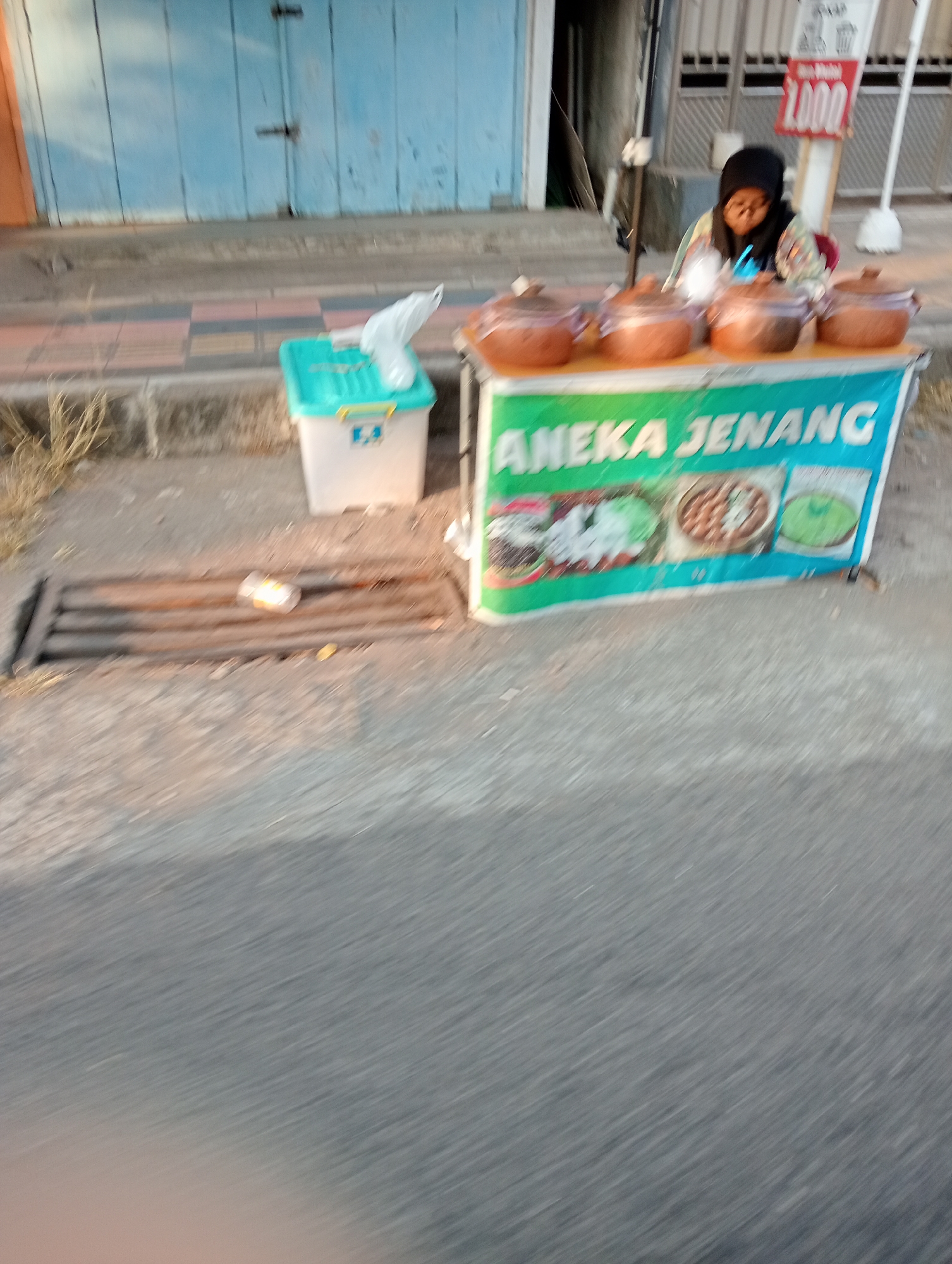 The image depicts an outdoor scene at a food stall in Grabag, Indonesia. A person wearing a black hijab and glasses is seated behind the stall, which displays several clay pots with lids, likely containing food. A banner on the front of the stall reads "ANEKA JENANG" and features images of various food items. To the left of the stall, a green and white plastic container is visible, along with some dried grass and a metal grate on the ground. In the background, a building with a blue door can be seen. A red sign with "1,000" is also visible on a pole. The scene appears to be captured from a moving vehicle due to the motion blur.