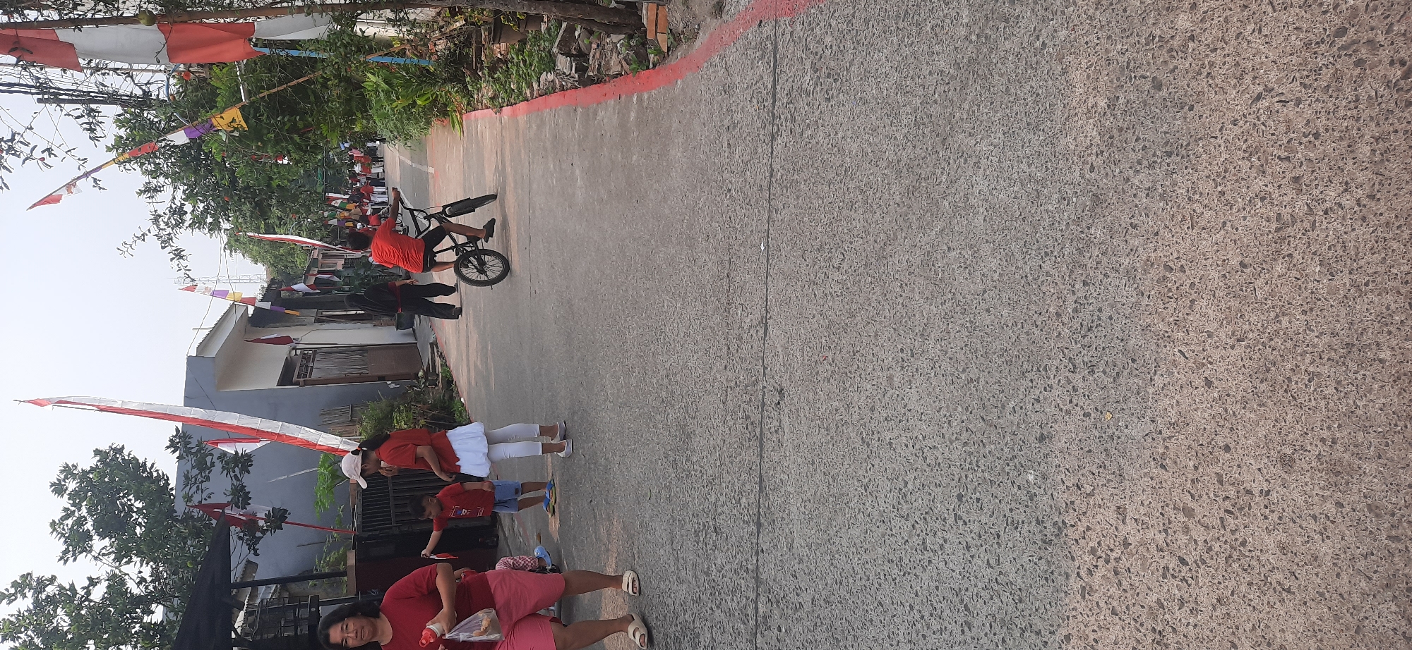 In Cisauk, Indonesia, a narrow paved road is lined with houses and trees. The road is decorated with Indonesian flags. In the foreground, a woman in a red shirt and pink shorts walks with a plastic bag and a red bottle. Beside her, a young boy in a red shirt and blue shorts holds a small flag. To the right, a child in a red shirt, white skirt, and white leggings stands. Further down the road, a boy in a red shirt rides a bicycle. Other people, some wearing red, are visible in the background. The sky is overcast.