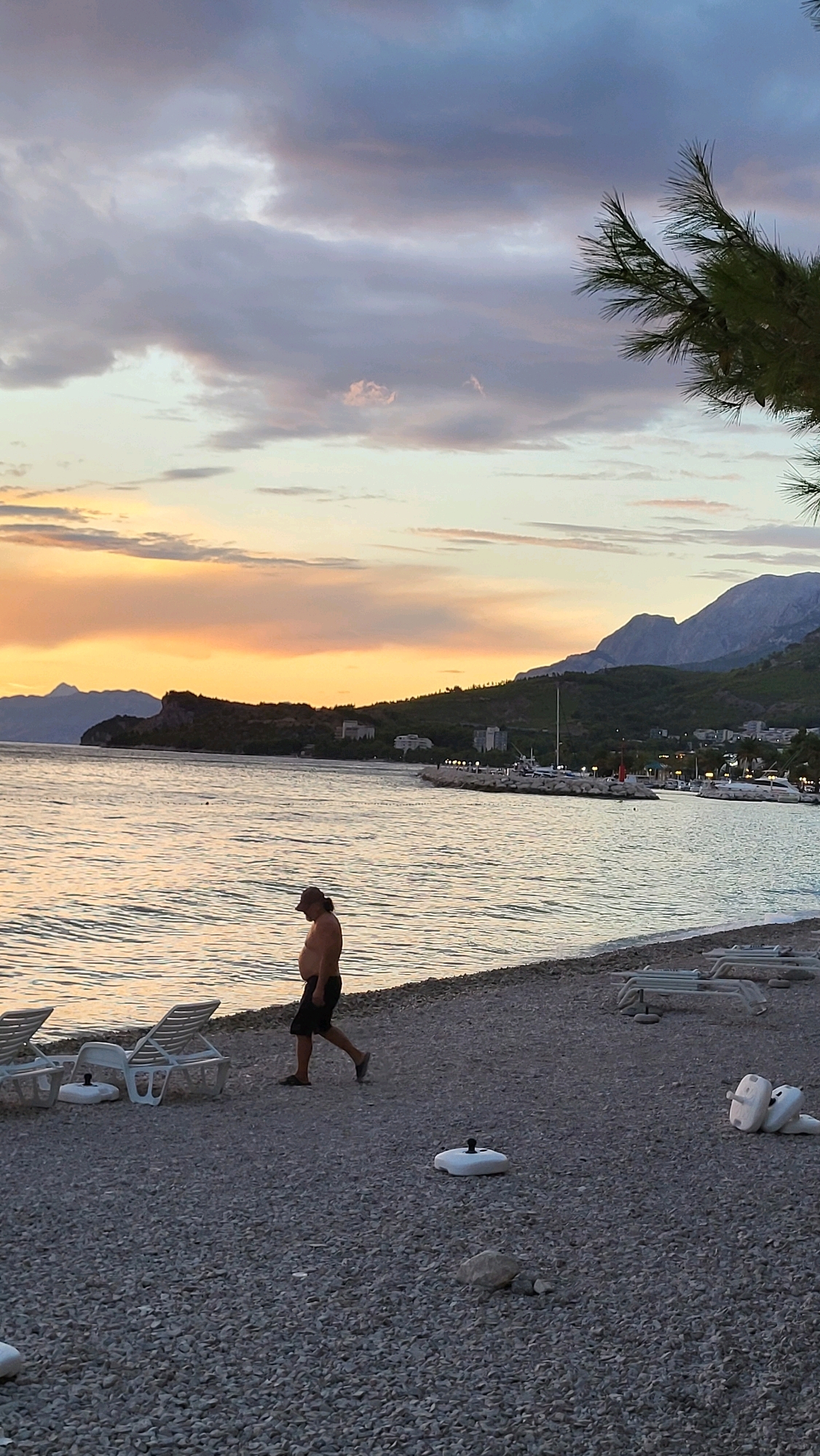 A lone, shirtless man wearing a baseball cap and shorts walks across a pebble beach. The sea is to his left, reflecting the warm hues of a sunset sky filled with clouds. In the distance, mountains rise behind a coastline dotted with buildings and a marina. The beach is lined with empty lounge chairs.